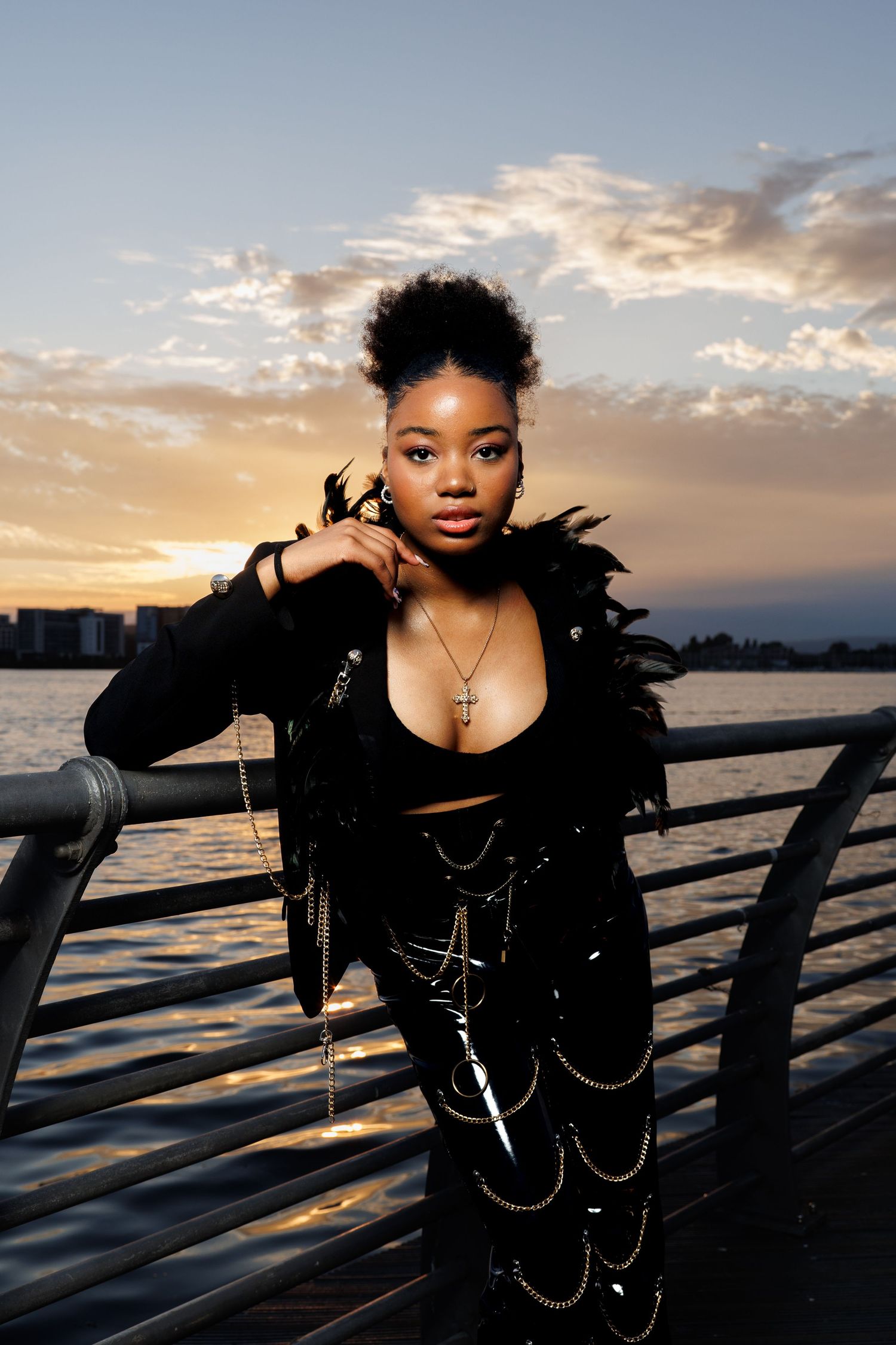 Young woman in black feathered jacket and chain-adorned pants posing on a pier at sunset with cross necklace.