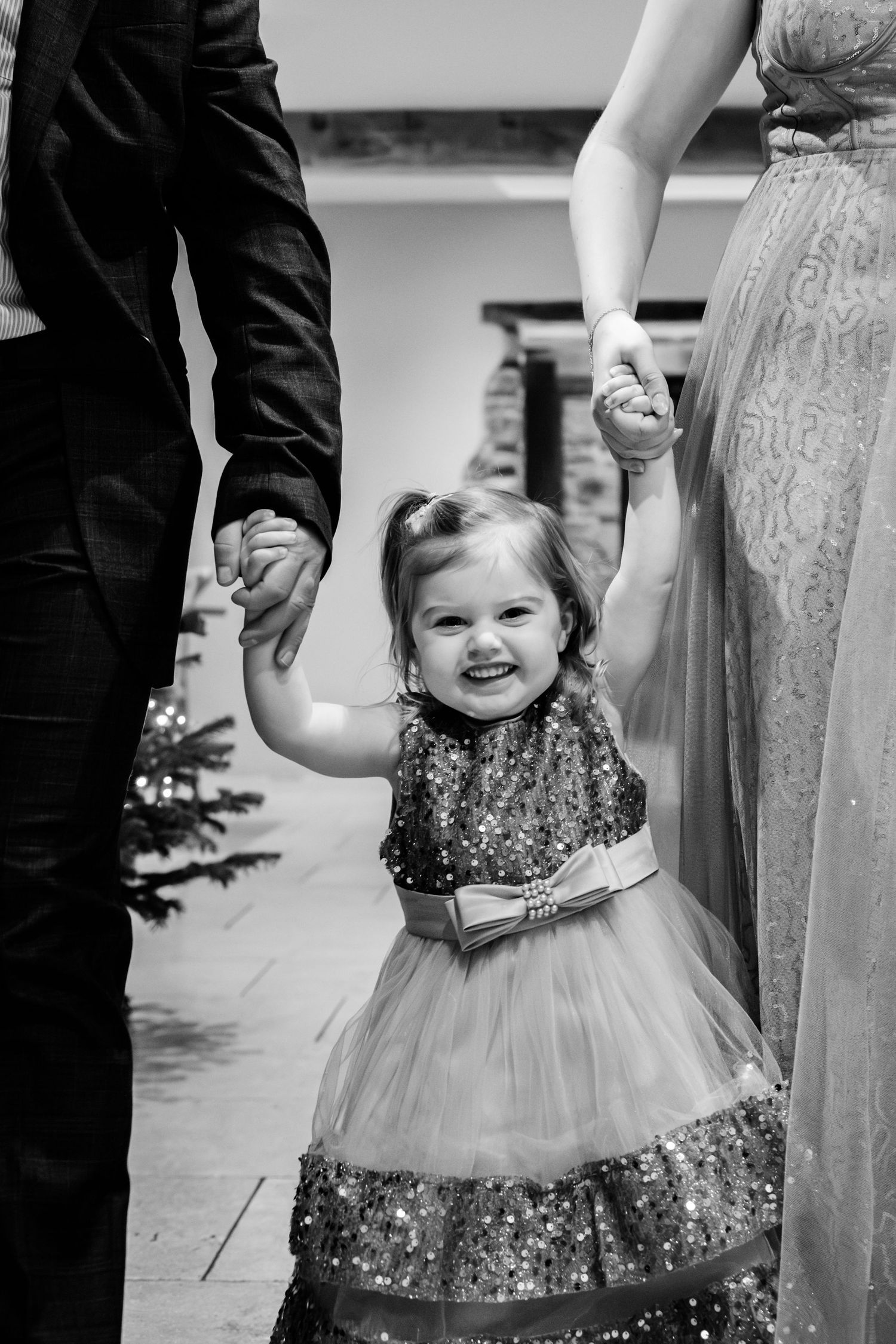 Black and white photo of a smiling toddler girl in a sparkly dress holding hands with her parents at a holiday event.