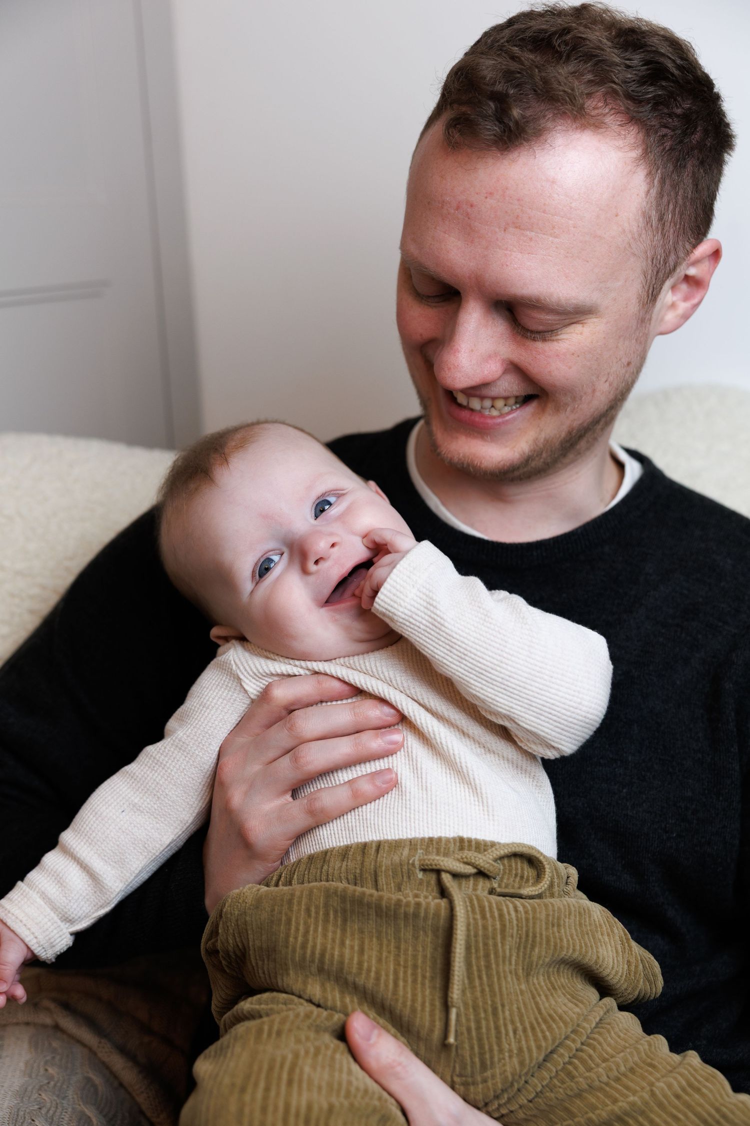 Smiling father holding his laughing baby dressed in cream top and olive pants on a cozy sofa.