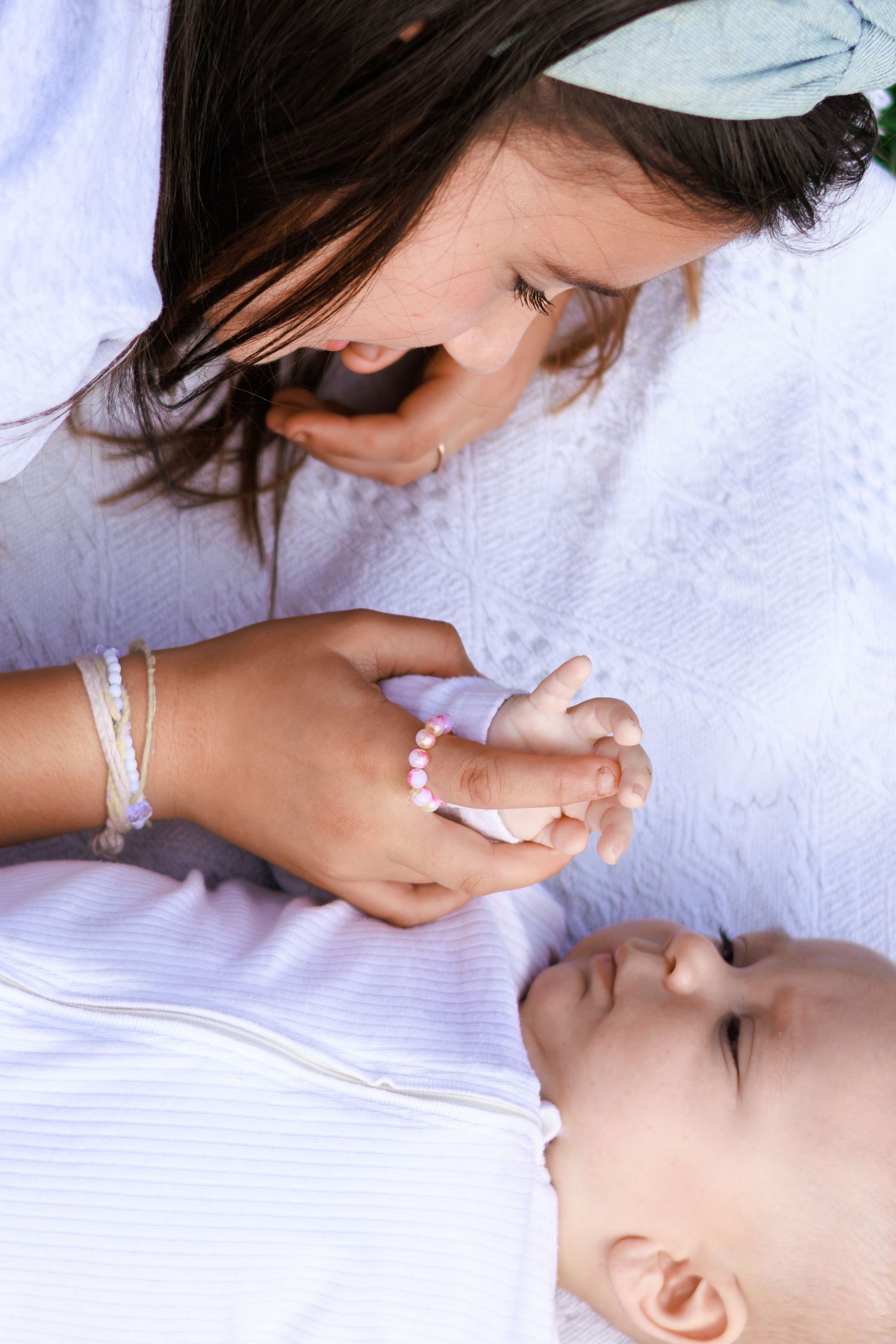 A young girl with bracelets tenderly holds a baby's hand while leaning close, both dressed in white.