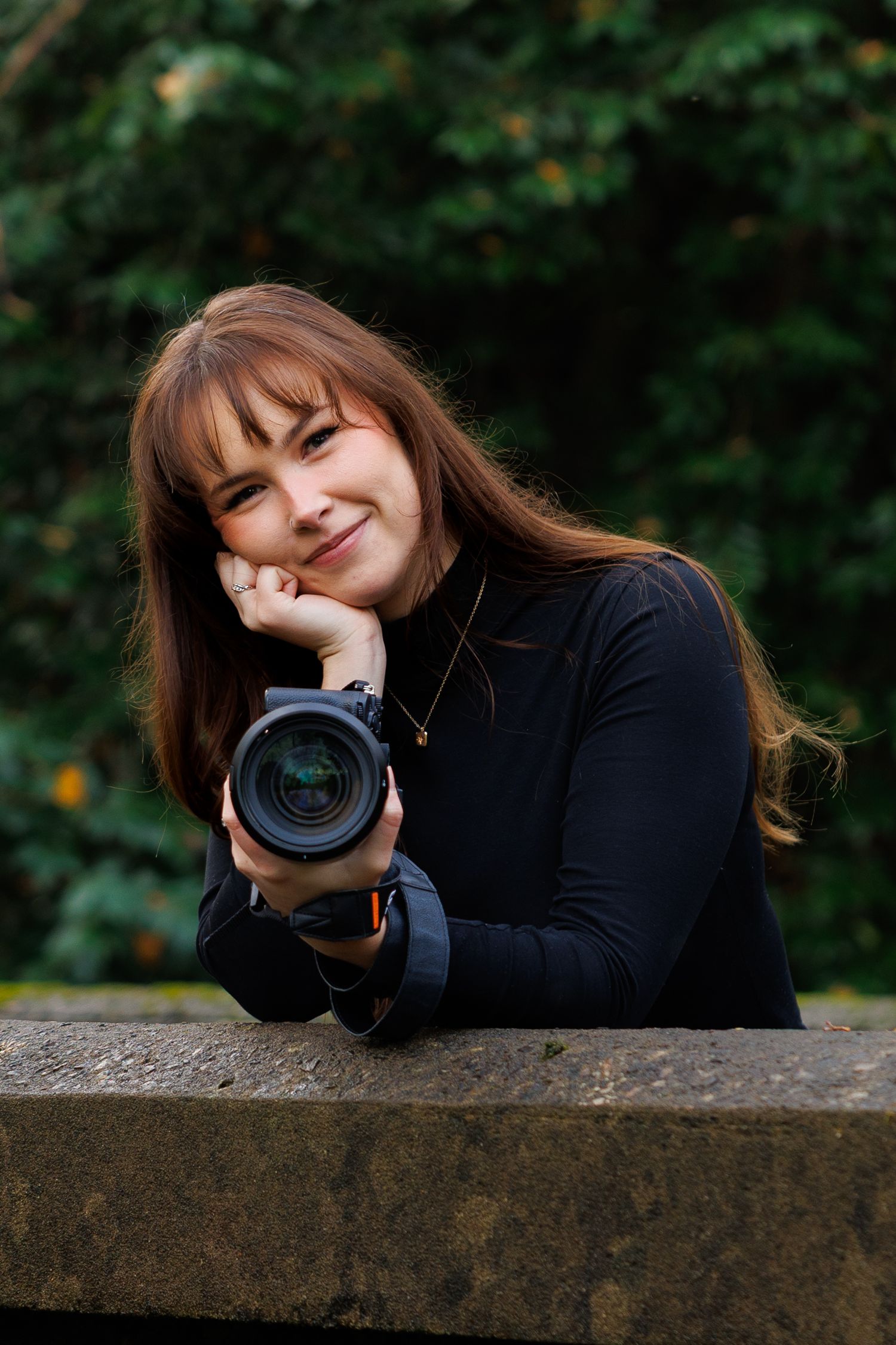 Smiling young female photographer resting chin on hand holding a camera in an outdoor setting.