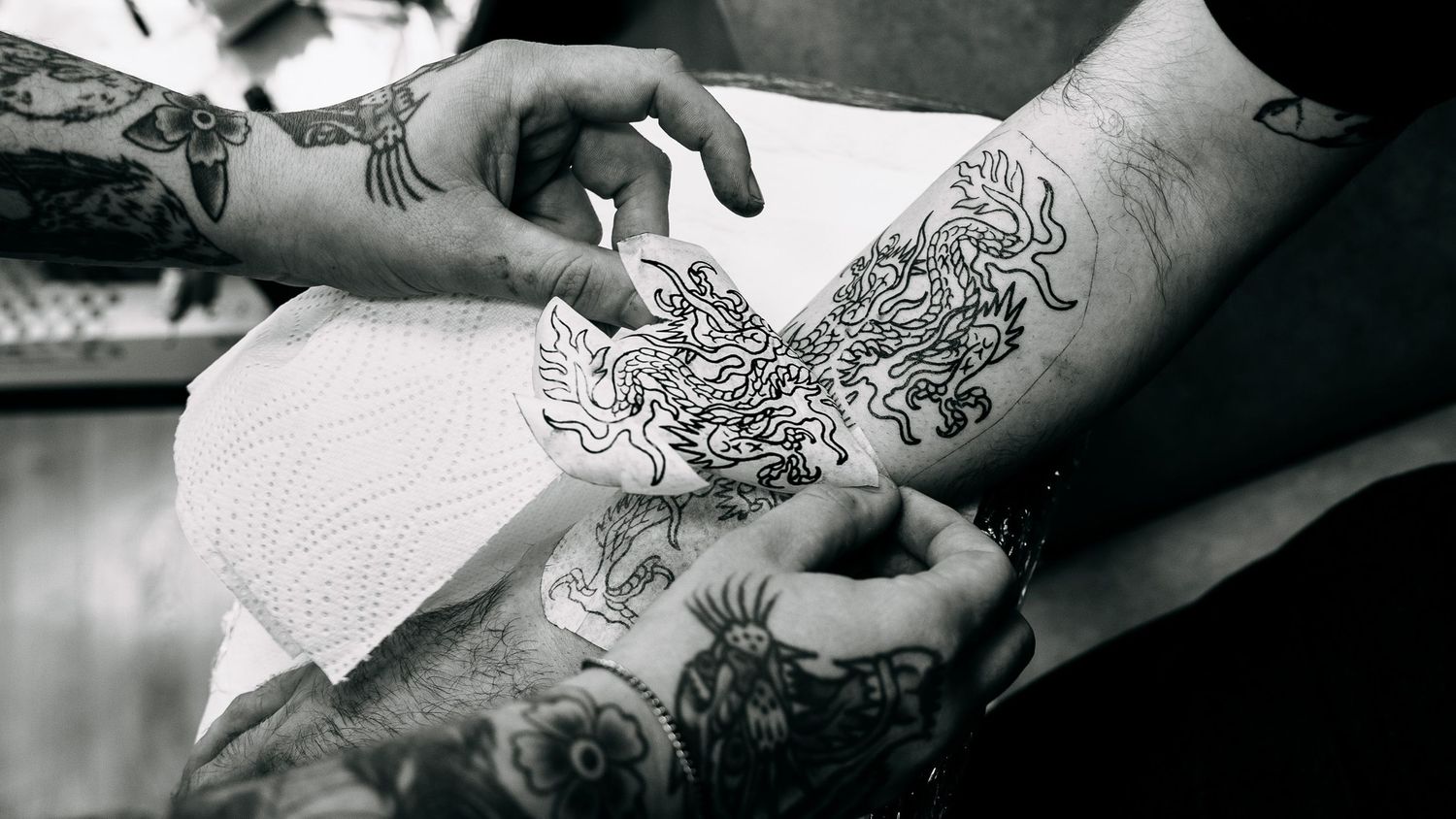 Black and white close-up of tattoo artist applying intricate dragon stencil to tattooed arm.