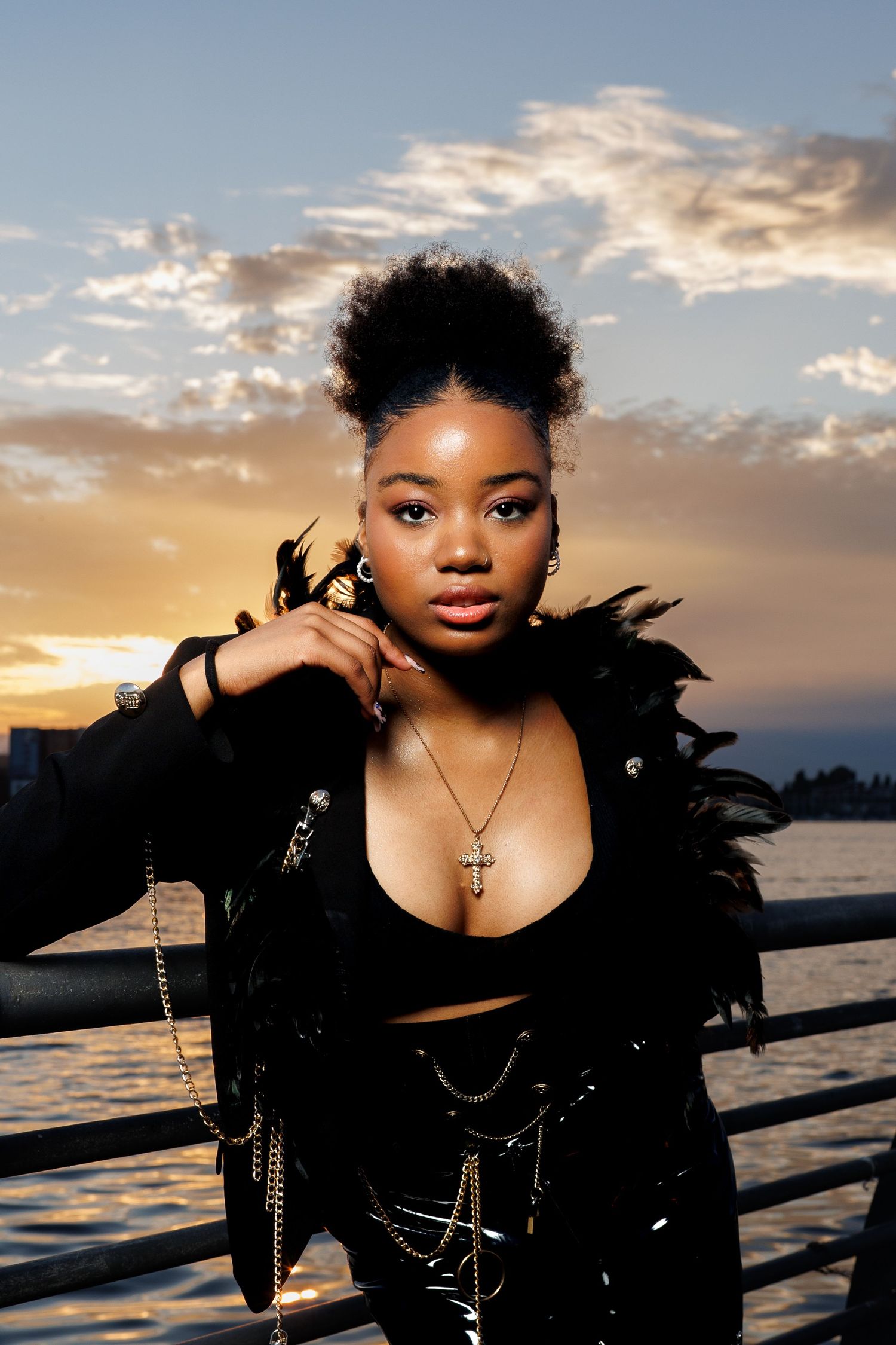 Young woman in black feather jacket with chain accessories posing at sunset waterfront pier.