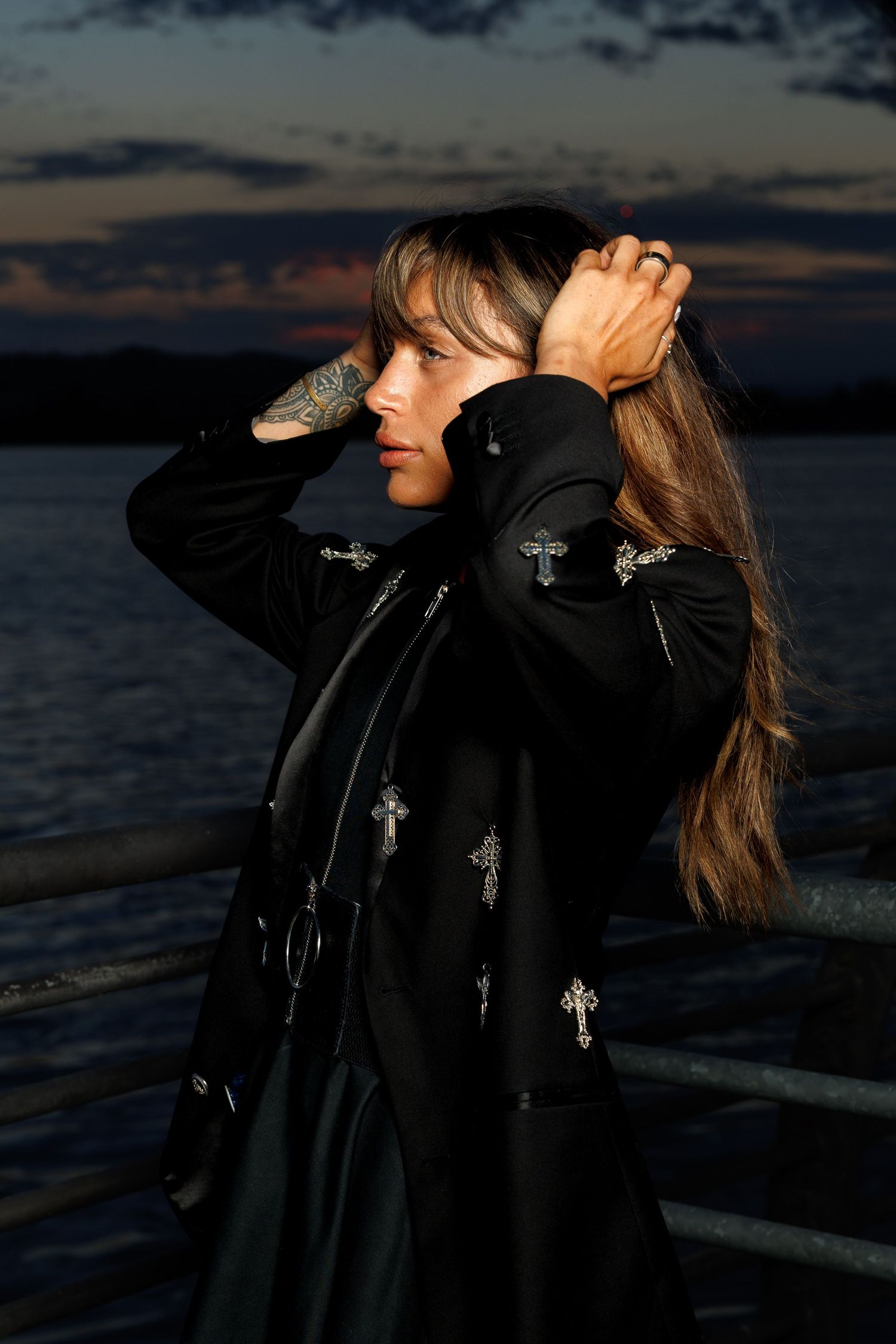 Young woman with long hair in black cross-embroidered coat poses by waterfront railing at dusk.