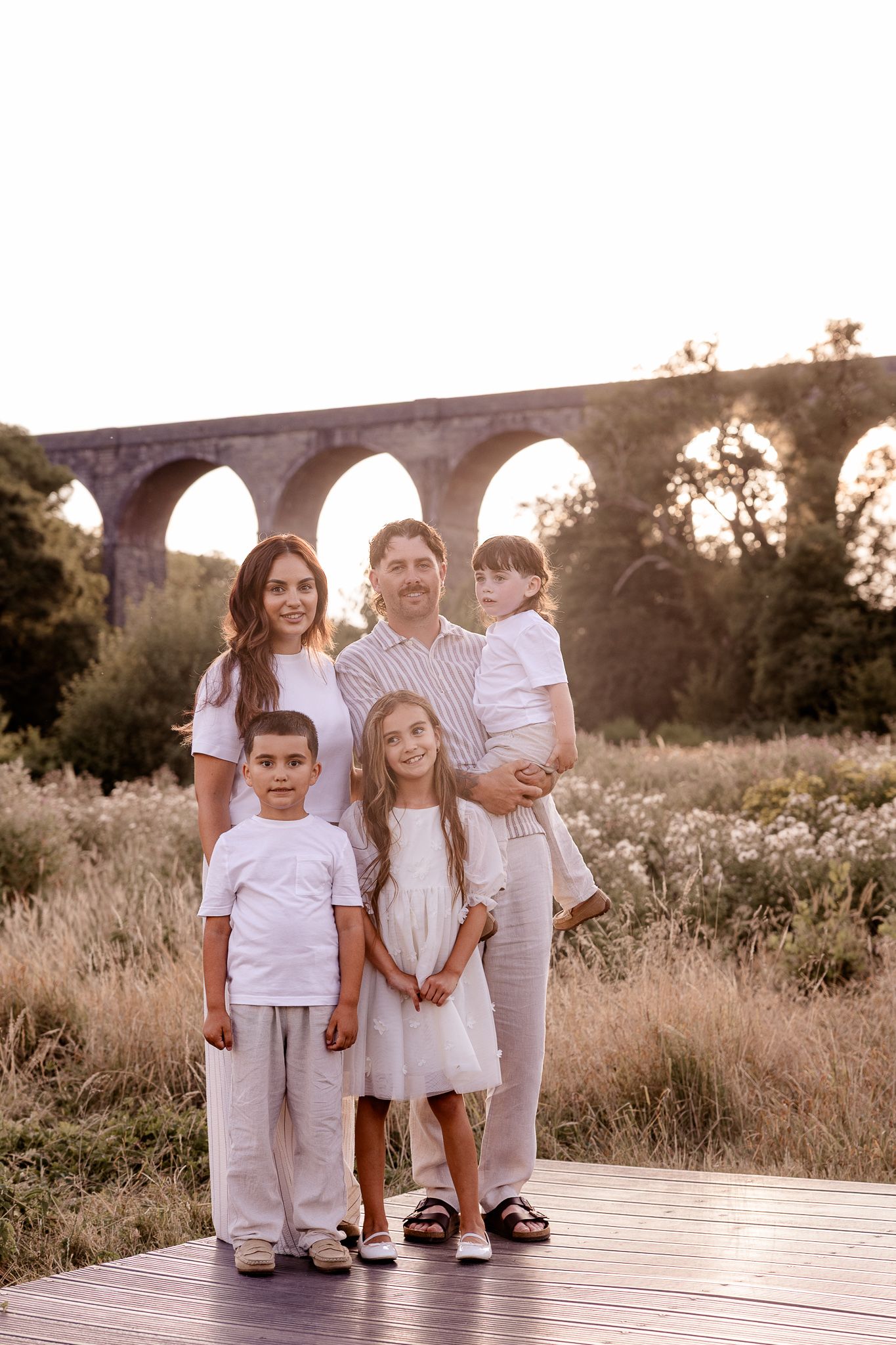 Family of five posing on wooden deck with historic stone aqueduct arches and golden meadow in background at sunset.