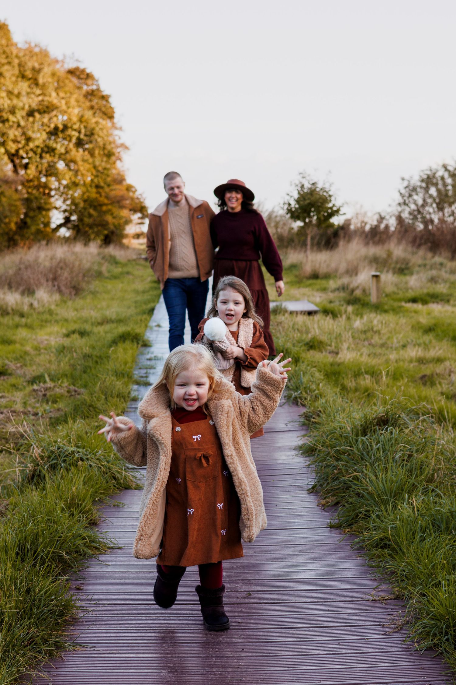 Family of four walking on a wooden boardwalk path through autumn fields, two young girls running ahead smiling.