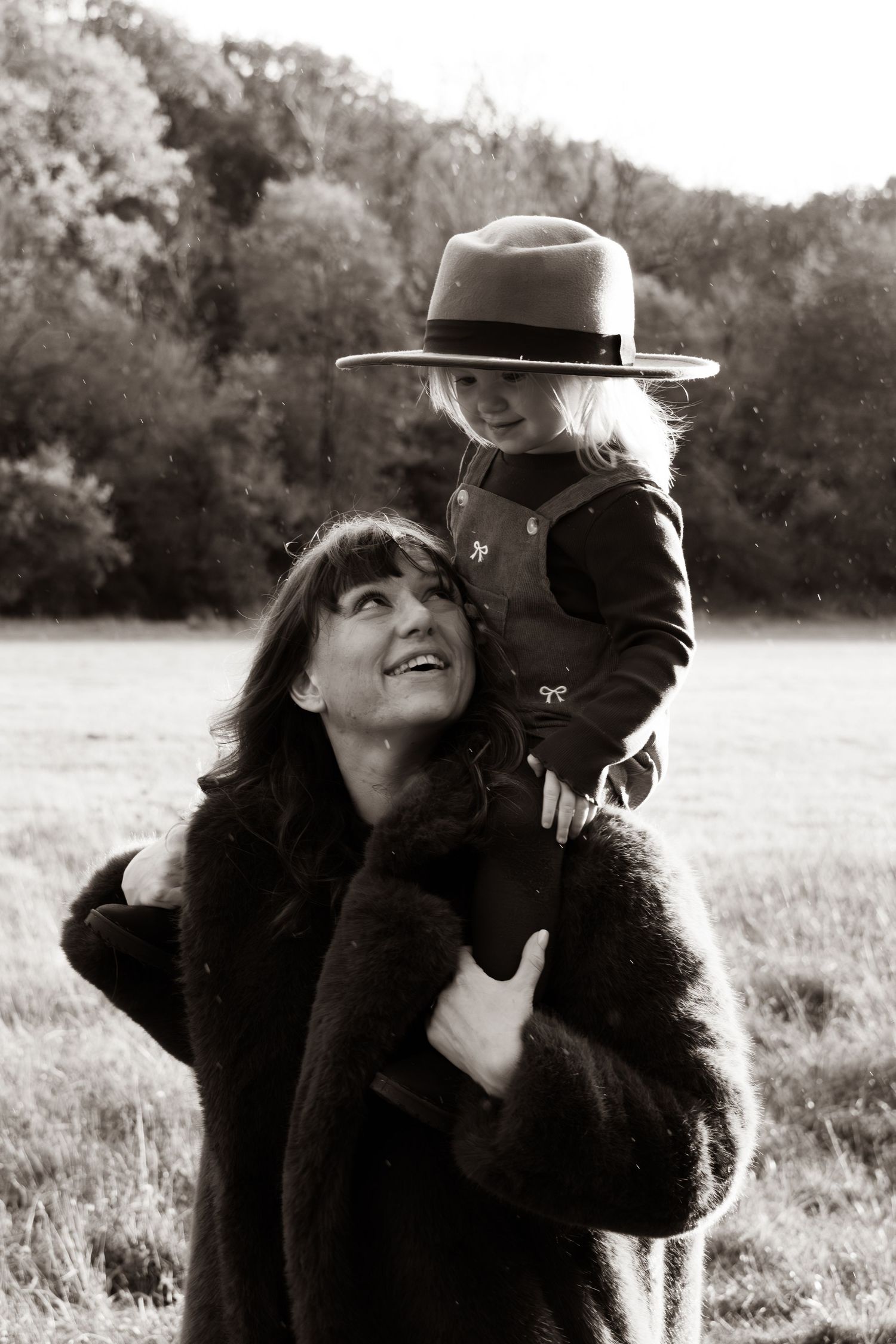 Black and white photo of mother carrying child in hat on shoulders in field.