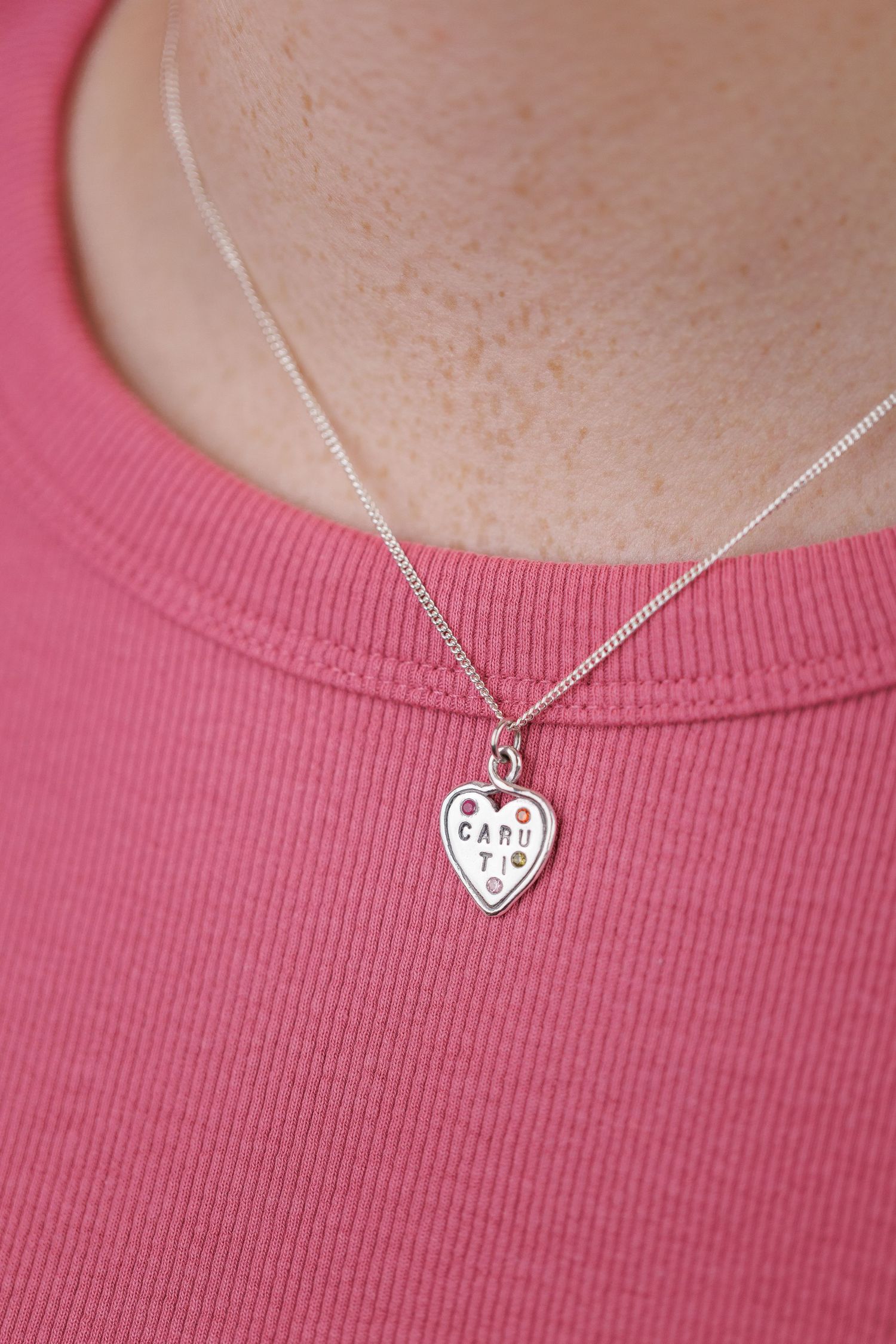 Silver heart charm necklace with CARU TI engraving worn on pink ribbed top.