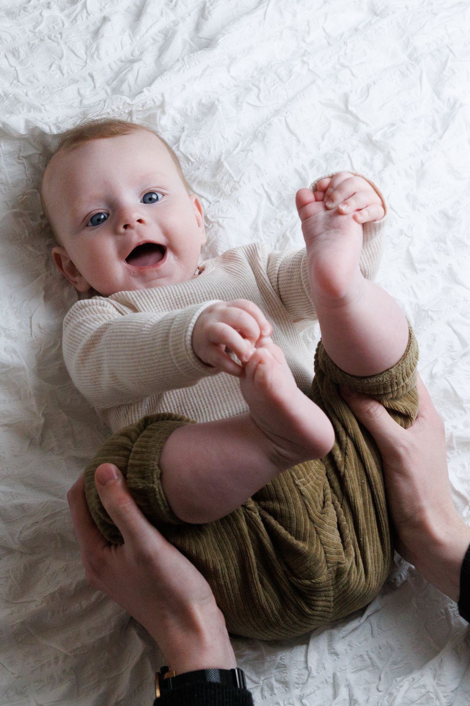 Happy baby with blue eyes laughing while lying on white blanket, wearing striped onesie and olive green pants.