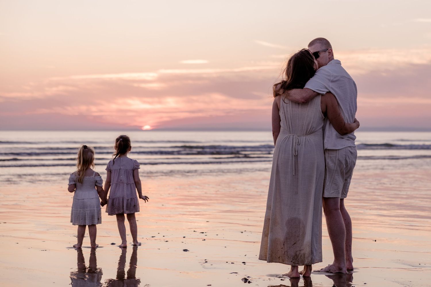 Family watching stunning pink sunset on beach, parents embracing while two young girls stand nearby.