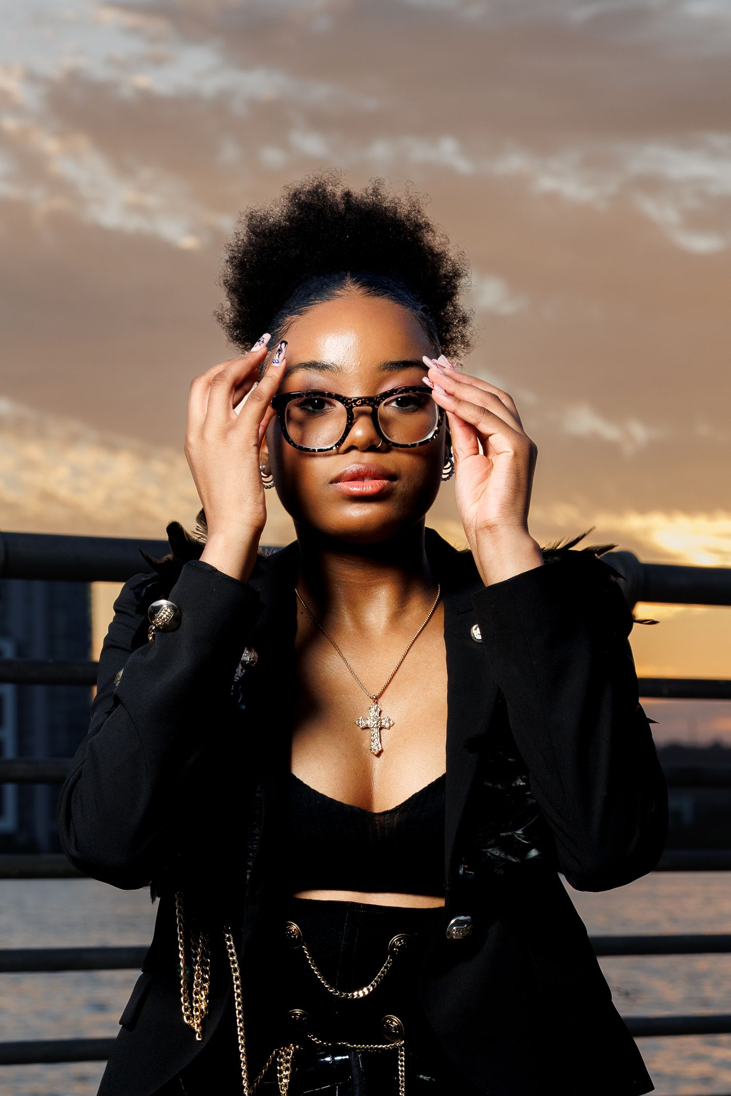 Young Black woman in black blazer adjusts oversized glasses at sunset, wearing cross necklace and chain accessories.