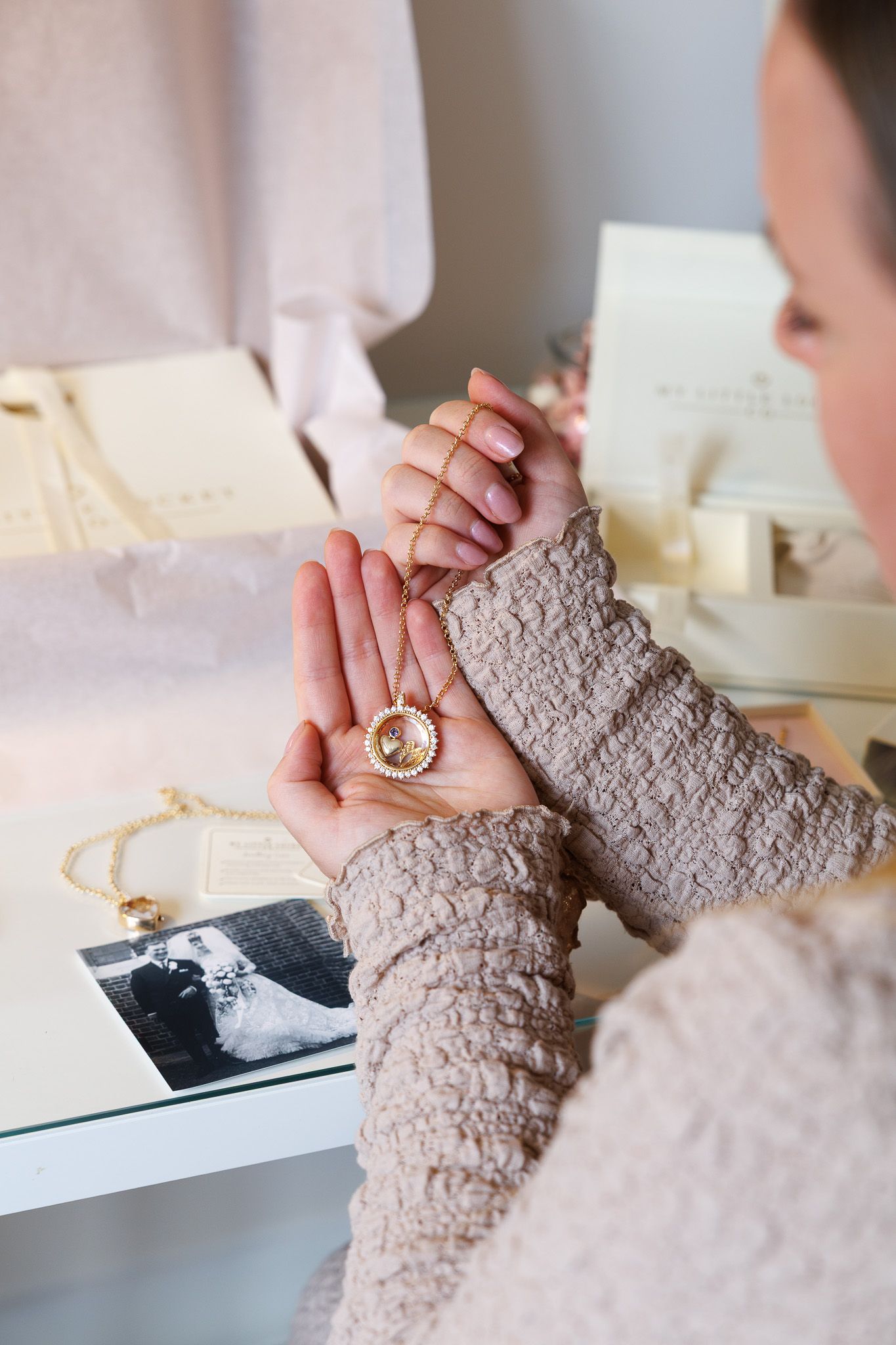 Woman holding a gold locket necklace over a photo, with jewelry and gift boxes on a glass table.