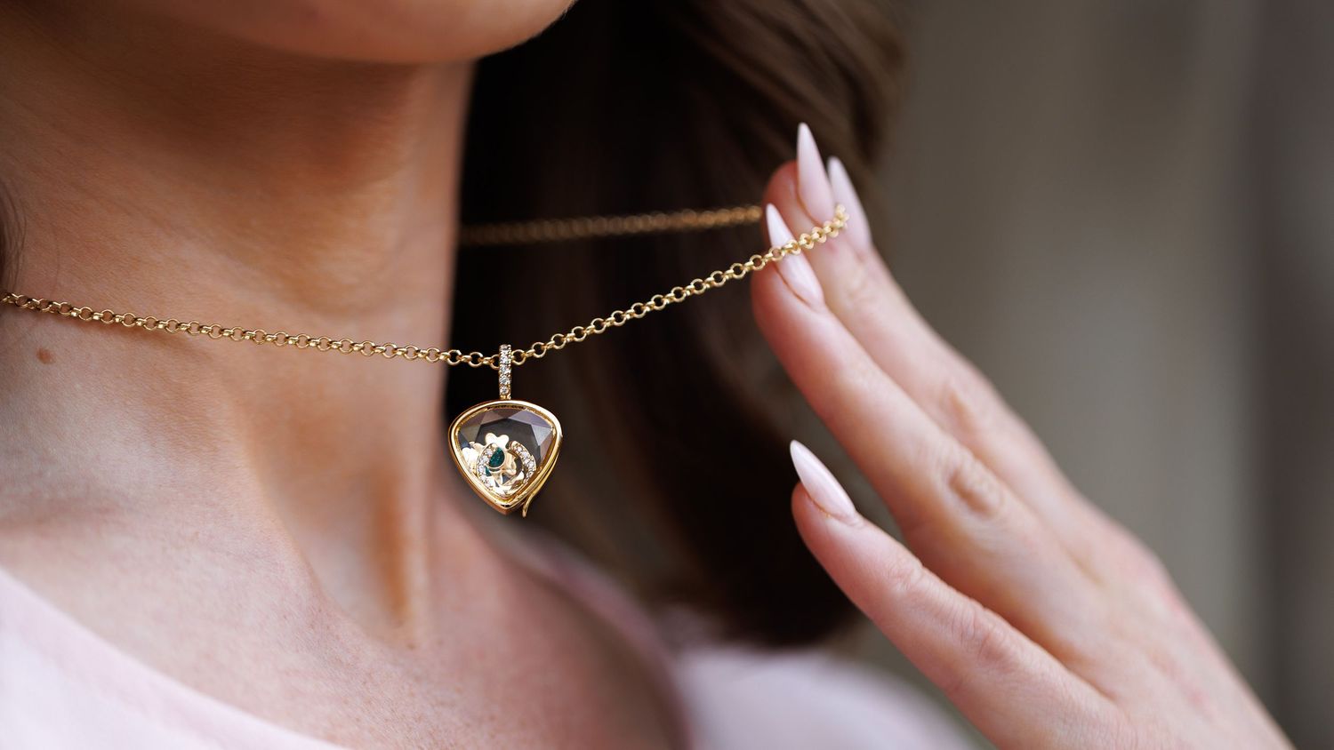 Woman wearing a gold heart-shaped crystal pendant necklace, touching it gently with manicured white nails.