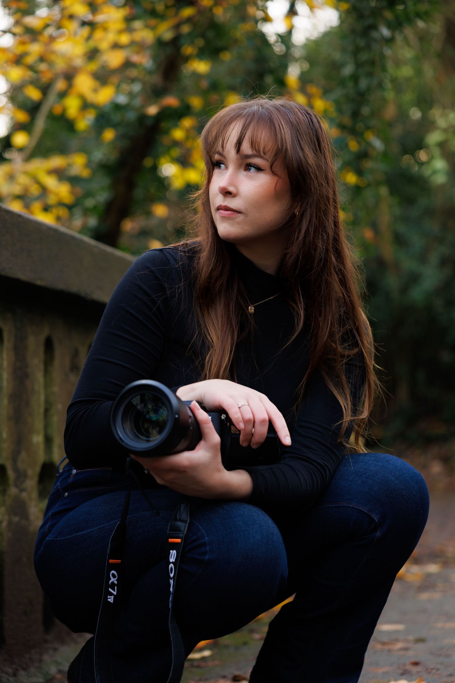 Female photographer crouching outdoors in autumn forest holding a Sony camera looking upward.