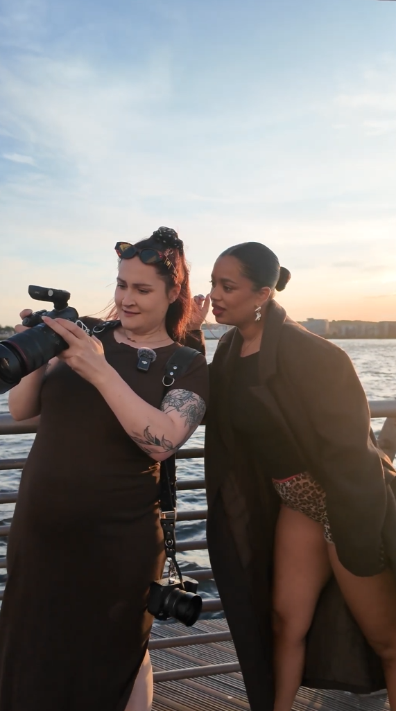 Two women review photos on a camera at a waterfront pier during golden hour sunset.
