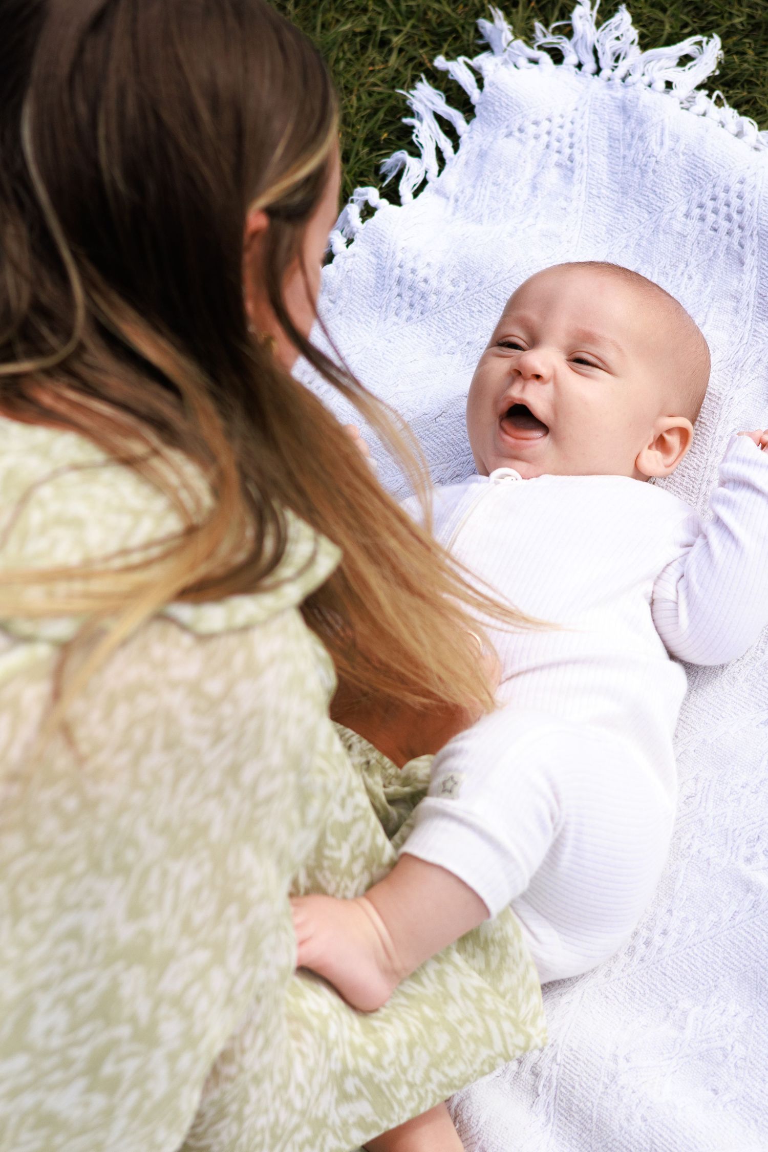 A happy baby in white onesie laughs on a white blanket outdoors while an older girl with long hair looks on.