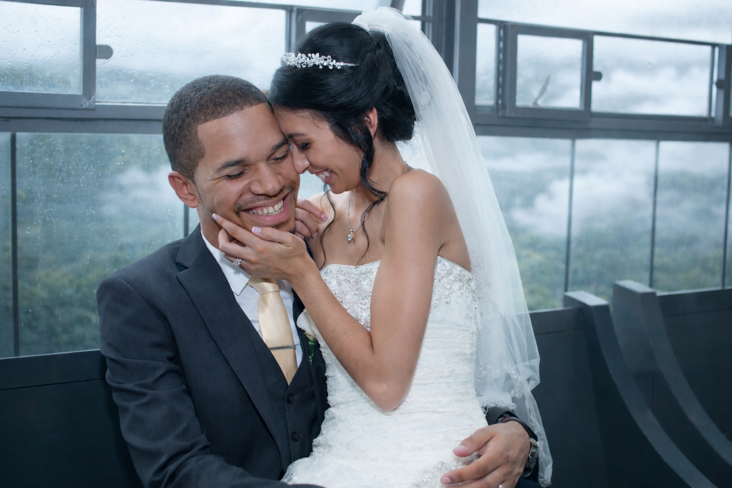 A tender moment between a bride and groom as they share a quiet laugh near a rain-covered window. Soft light and natural emotion create a ti