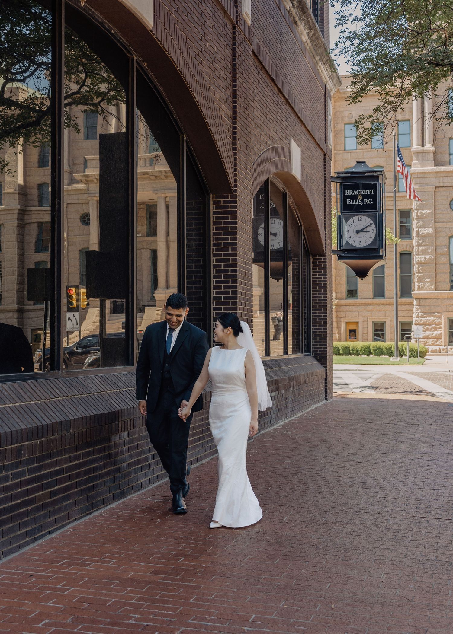 Bride and groom walk hand in hand on a brick sidewalk outside a historic building on their wedding day.