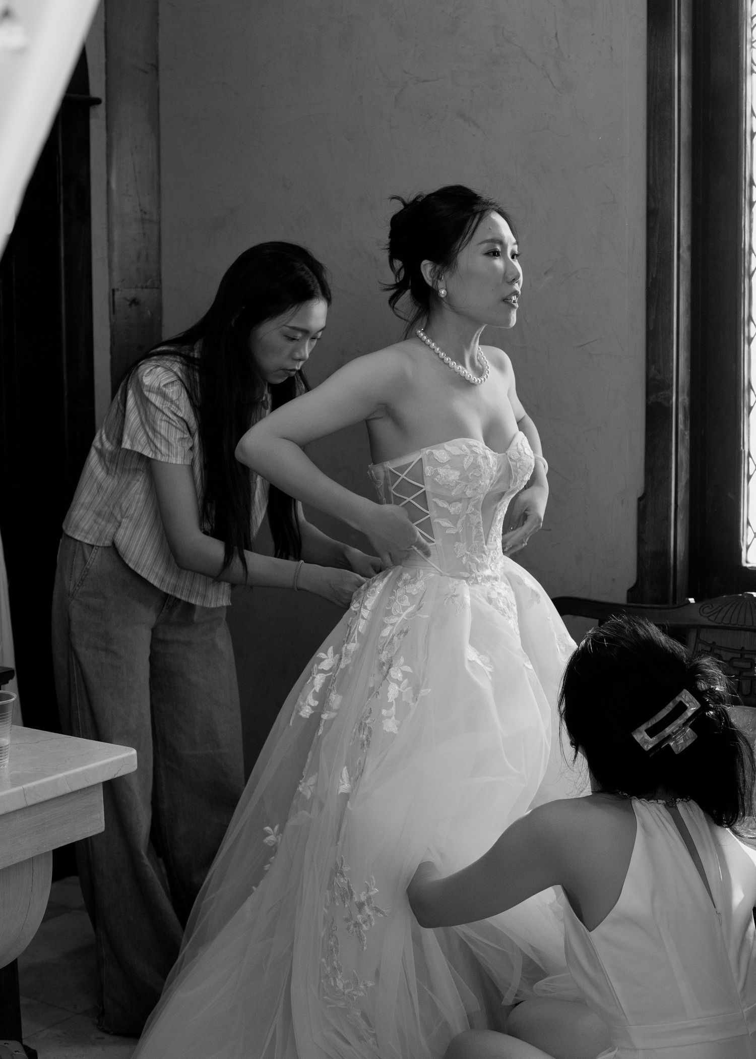 Bride being helped into a strapless lace wedding gown by two assistants in a black and white photo.