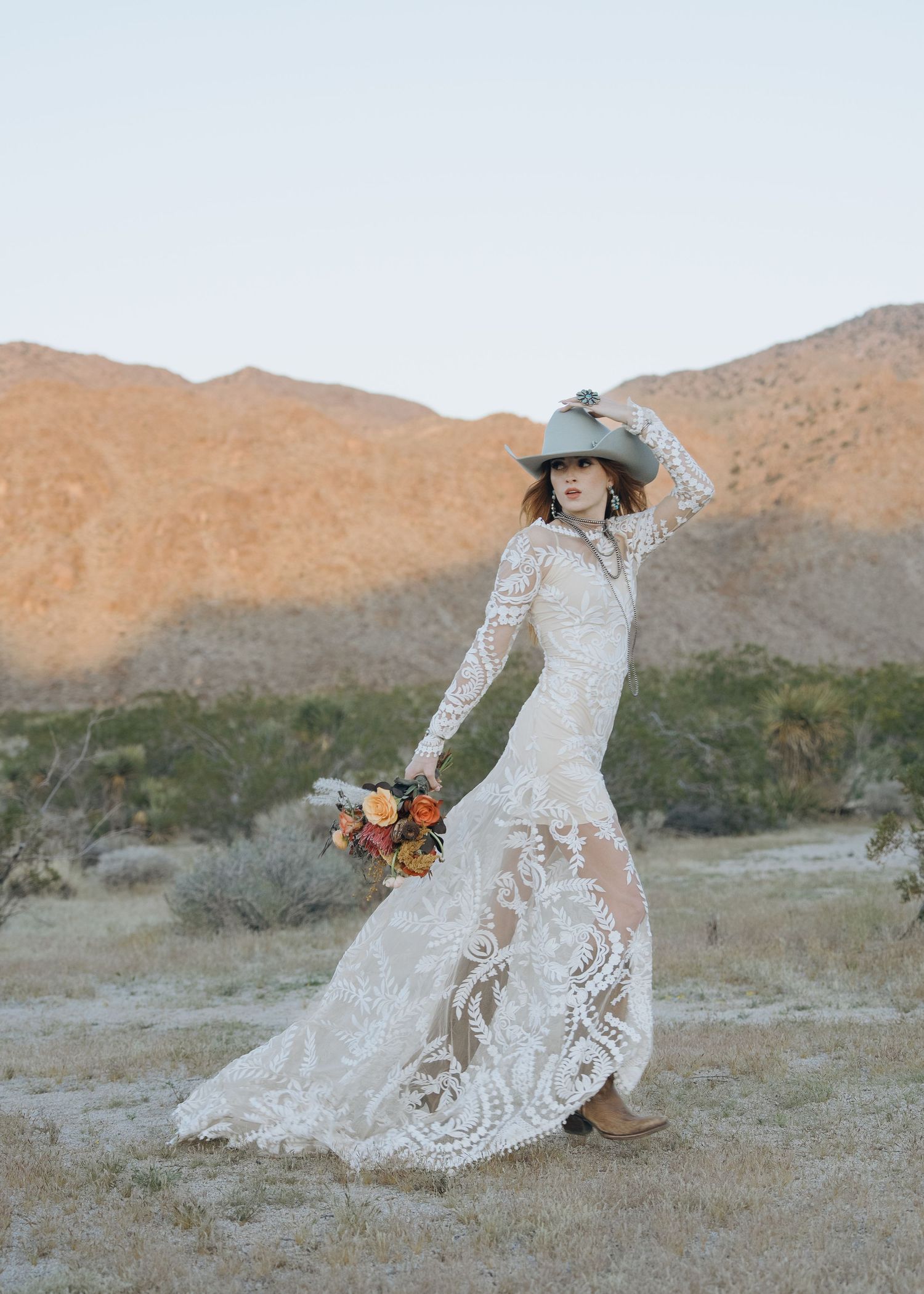 Western bride in lace mermaid gown and cowboy hat holding bouquet with mountain backdrop.