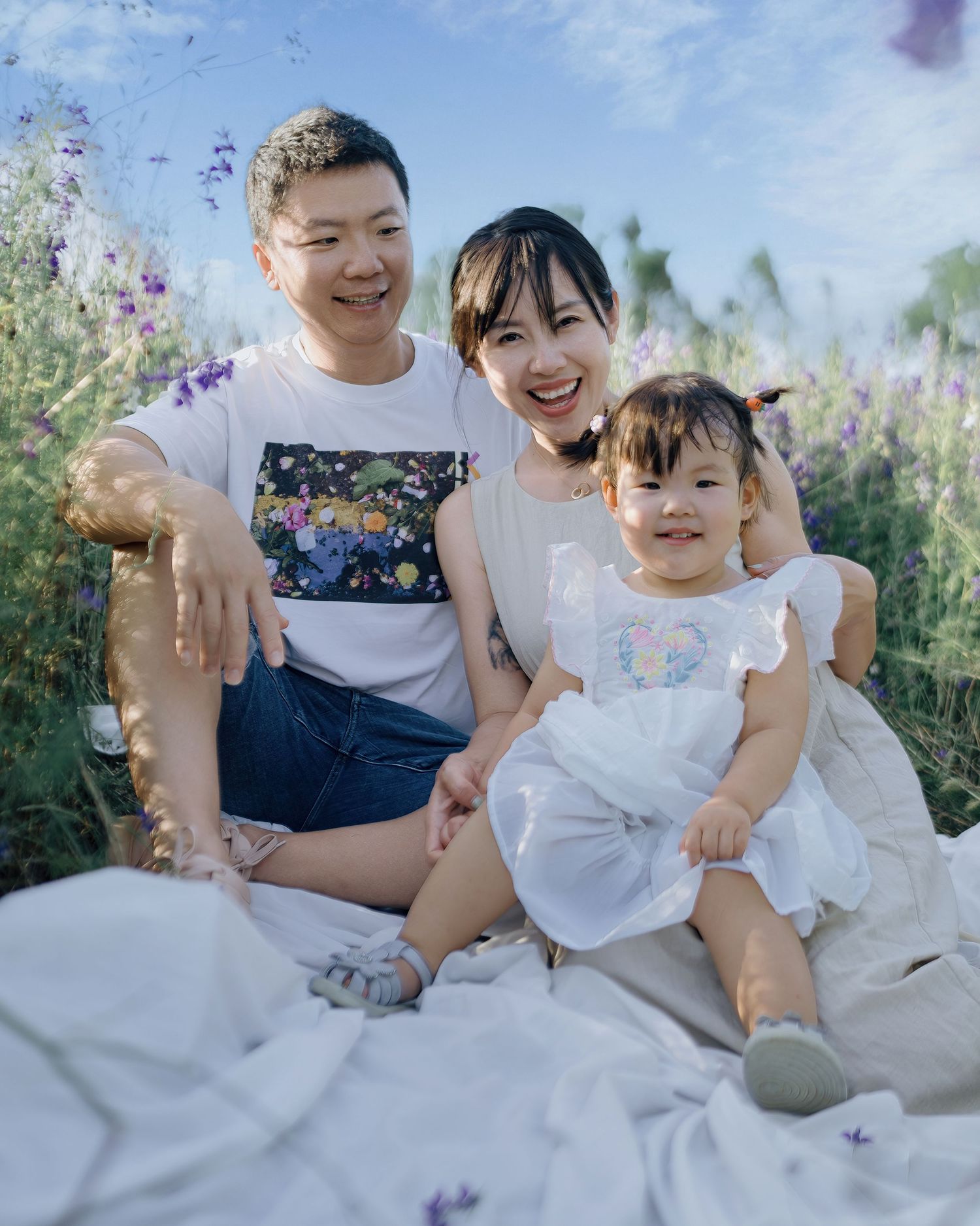A happy Asian family of three sits together on a white blanket in a purple wildflower field.
