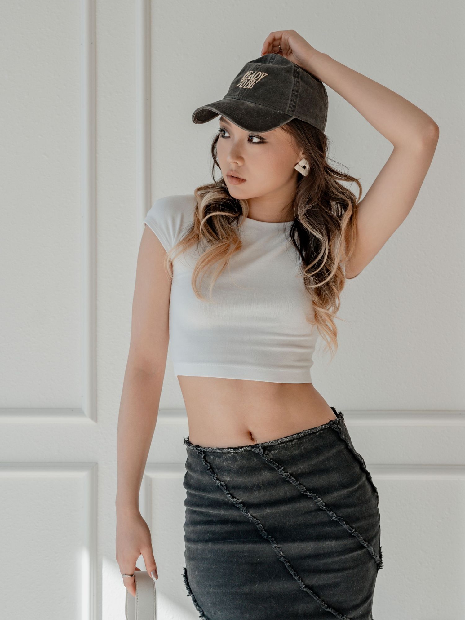 Stylish young woman in white crop top and dark denim skirt posing with baseball cap indoors.