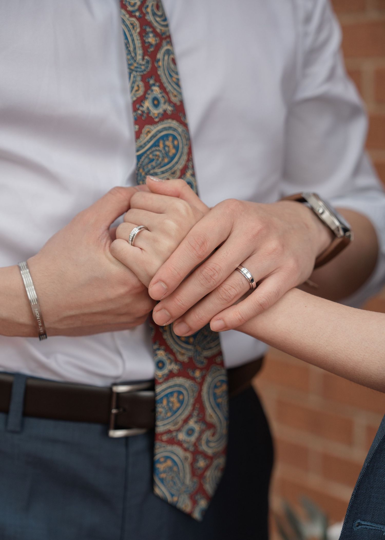 Close-up of newlyweds holding hands showing matching wedding bands, groom wearing paisley tie.