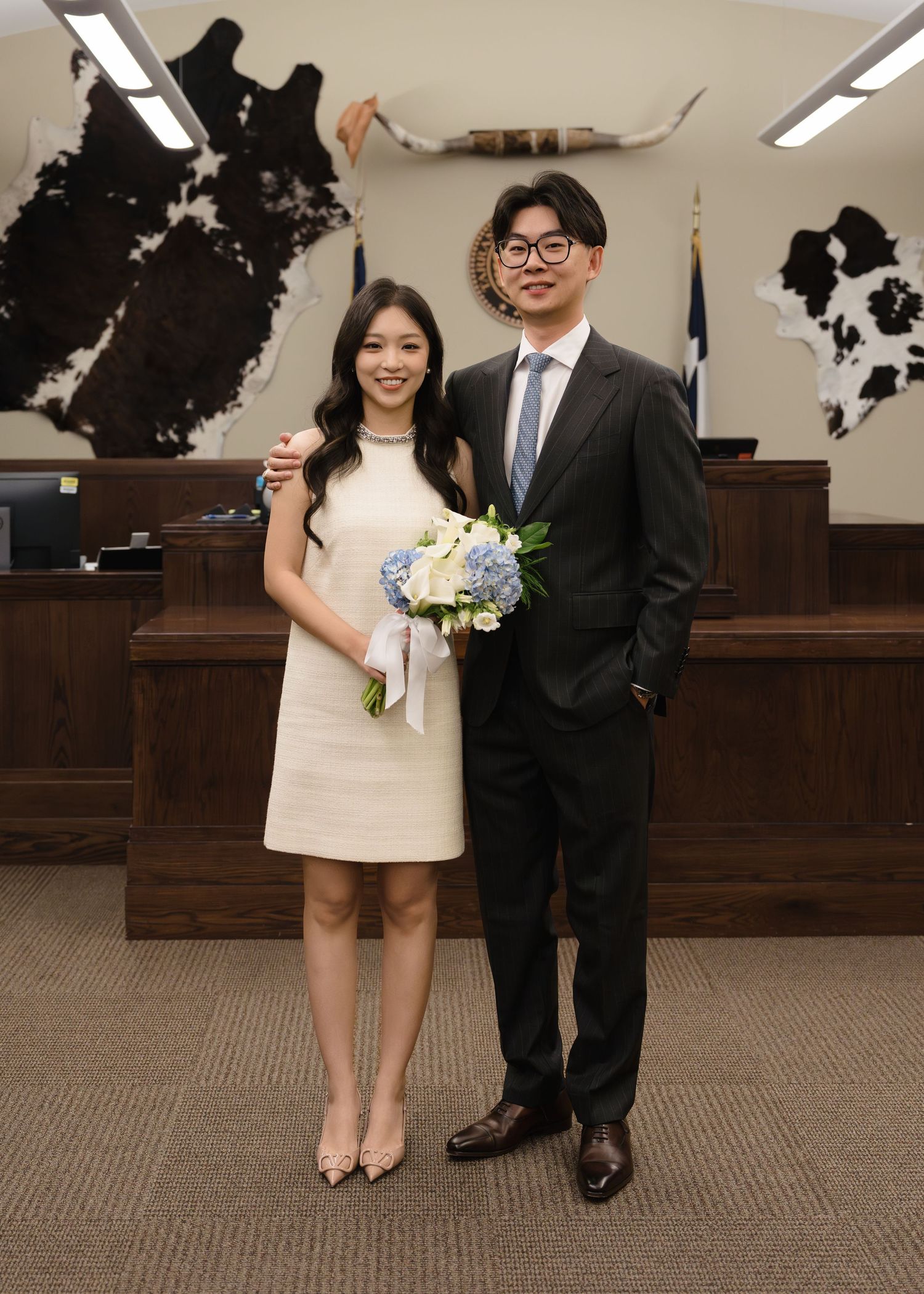 Asian couple posing in courthouse with longhorn decor, bride in white dress holding blue bouquet.