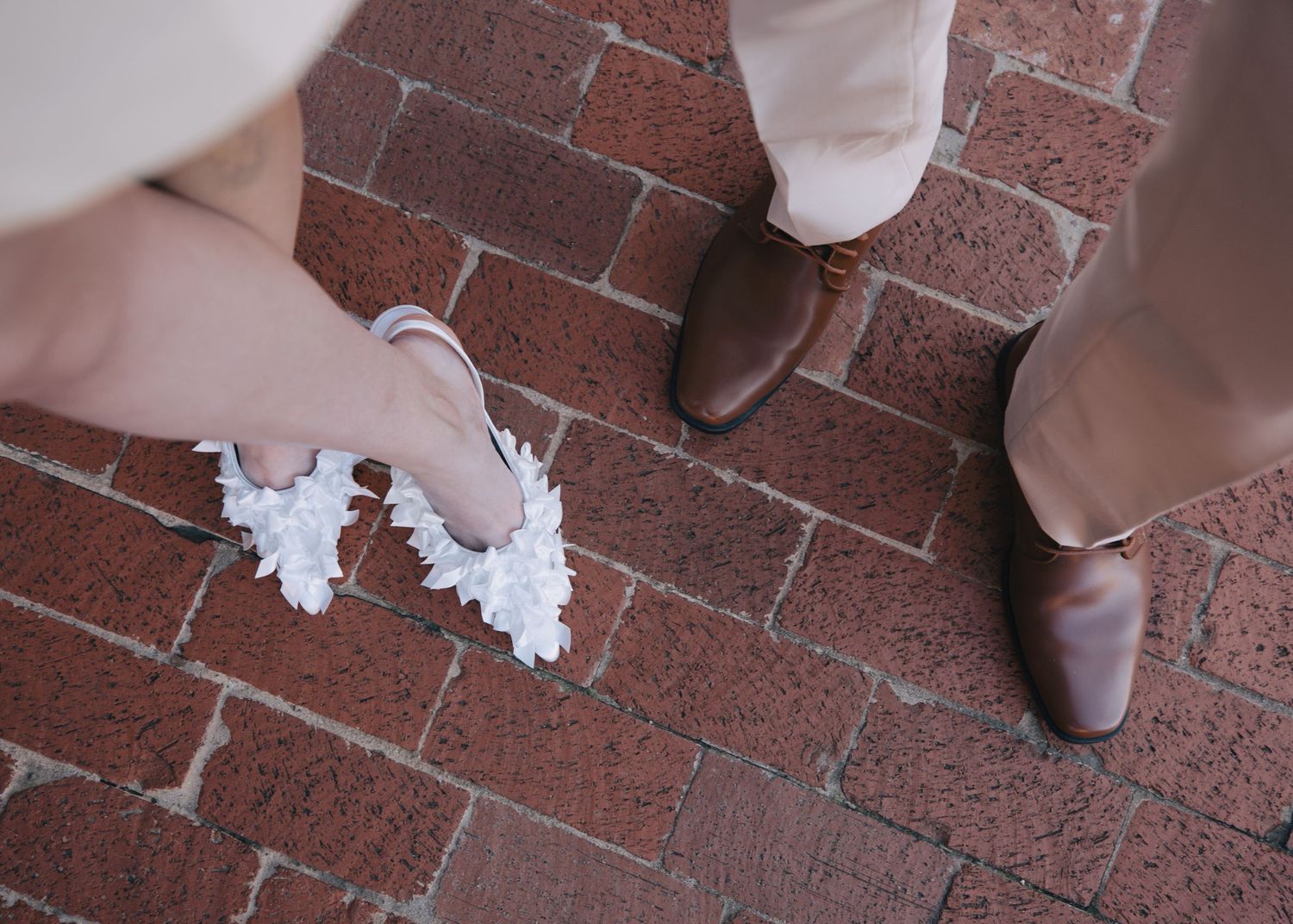 Close-up of bride's white floral heels and groom's brown leather dress shoes on red brick pavement.