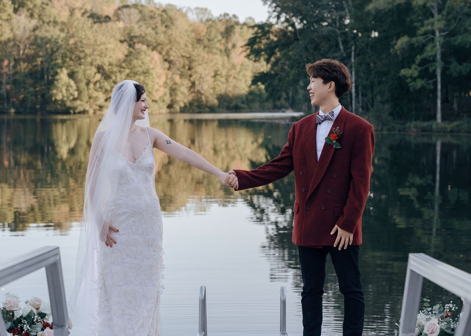 Bride and groom hold hands on lakeside dock, she in lace gown with veil, he in burgundy suit.