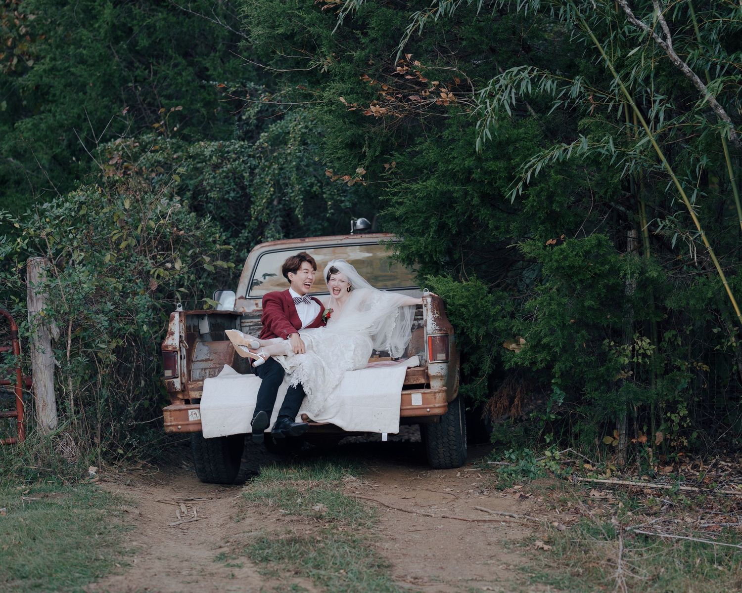 Bride and groom sitting together in bed of rustic pickup truck on wooded dirt road.