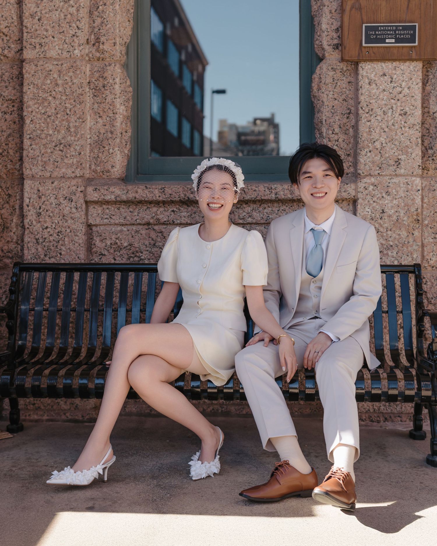 Smiling couple in wedding attire sits on a bench outside a historic brick building on a sunny day.