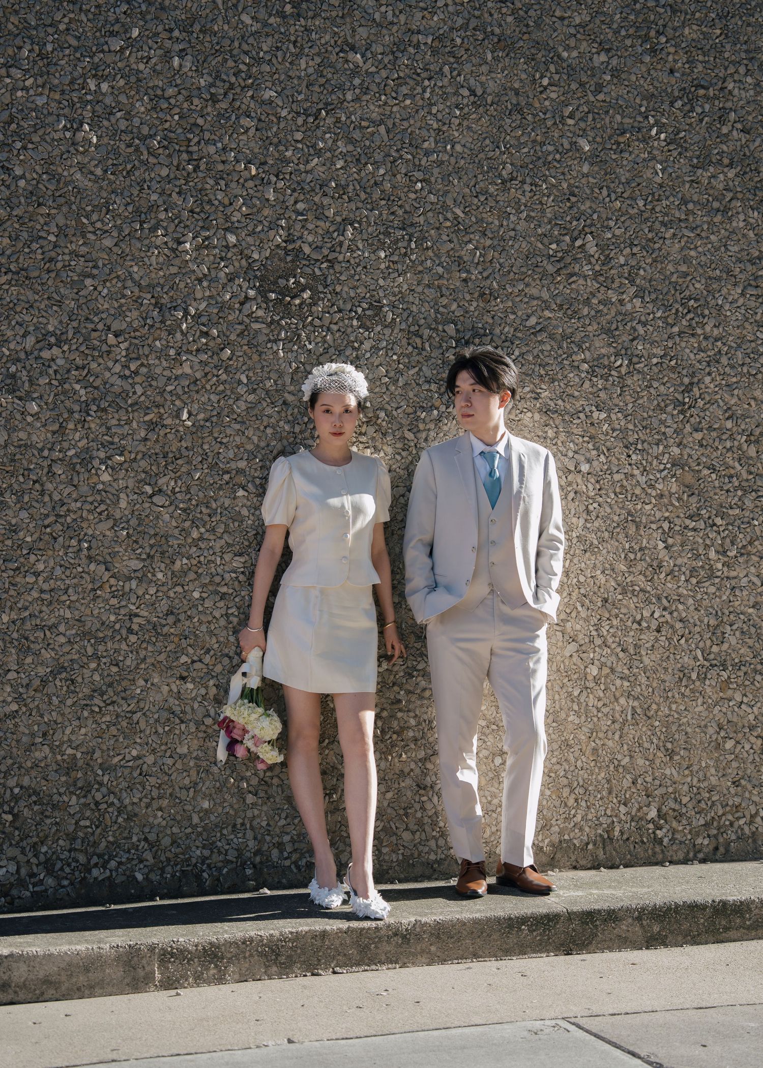 Bride in white mini dress and groom in beige suit posing against a textured concrete wall.