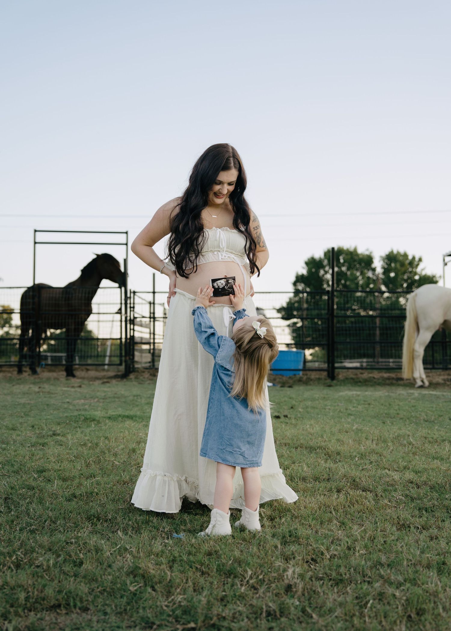 Pregnant woman in white dress with young daughter holding ultrasound photo on a farm with horses.