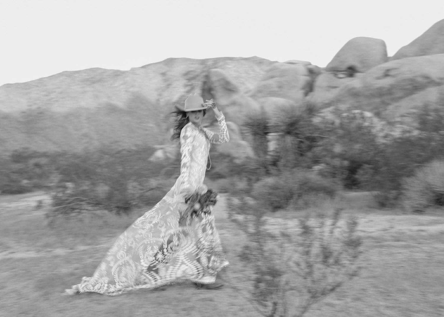 Artistic black and white photo of bride in lace gown walking through desert landscape.