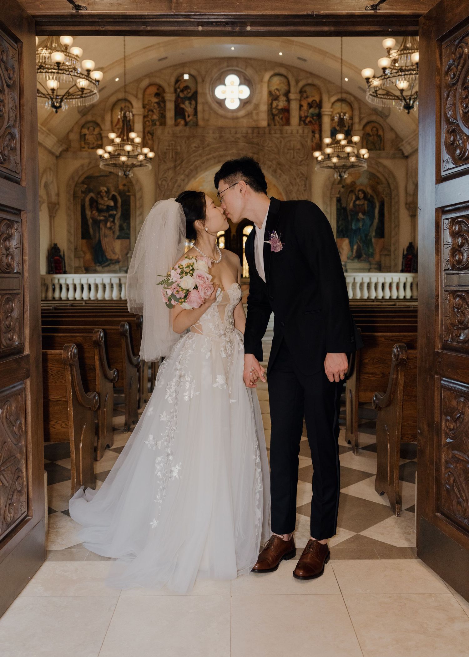 Bride and groom share a kiss at the chapel altar, bride in floral lace gown holding bouquet.