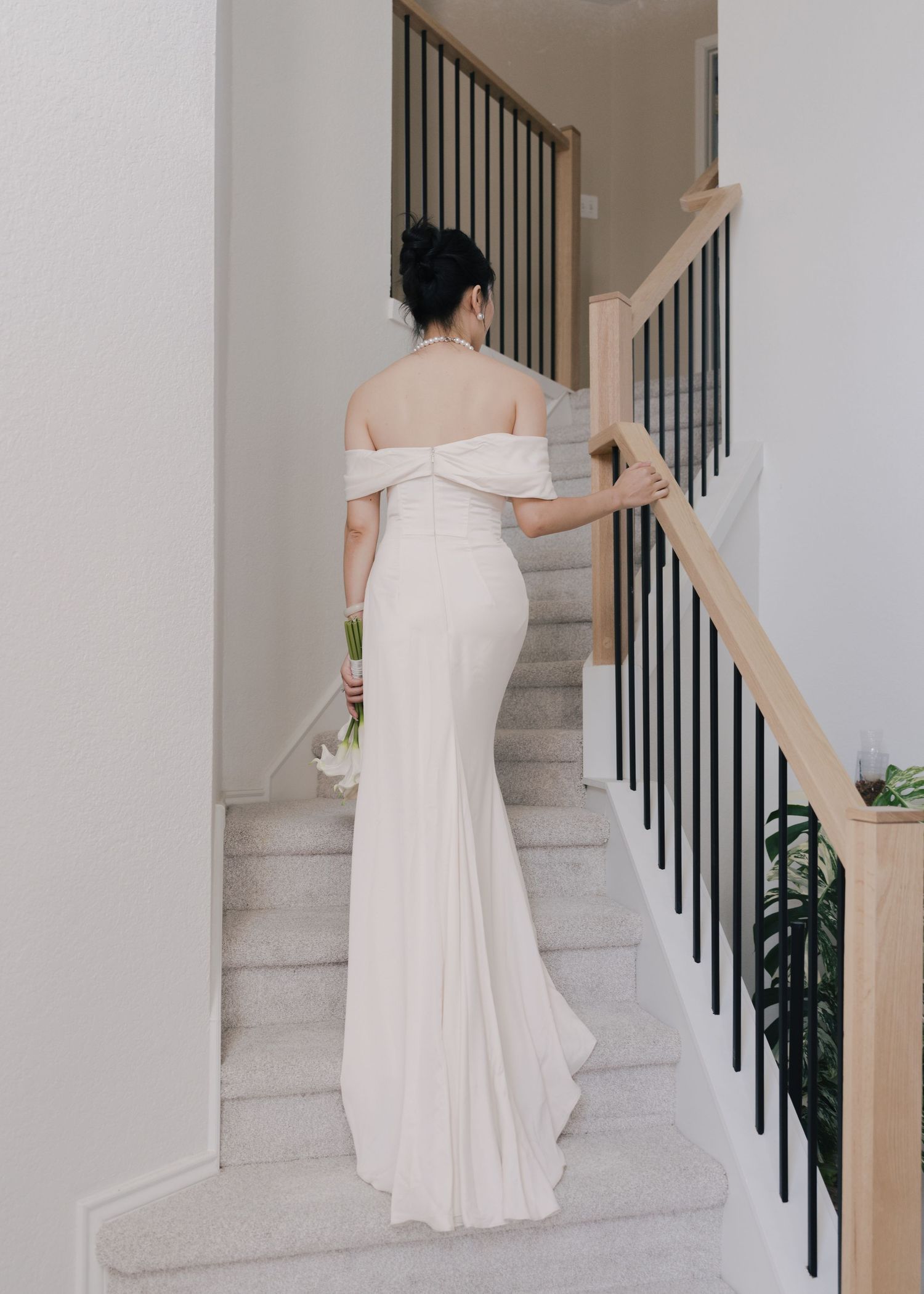 Bride in elegant off-shoulder white gown walks up modern staircase holding bouquet.