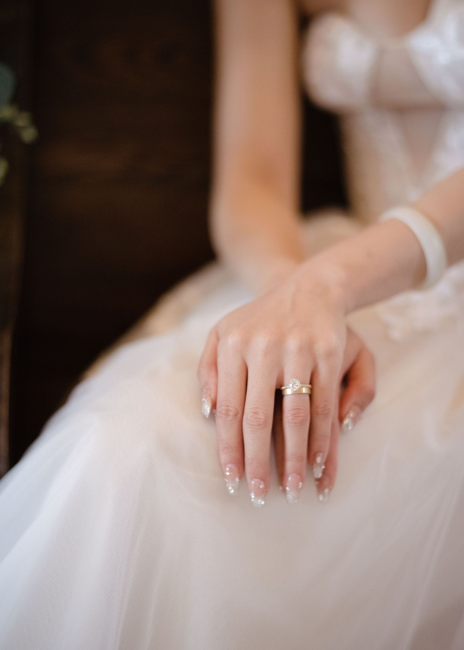 Close-up of bride's hands in white tulle gown showcasing a delicate gold engagement ring with manicured nails.