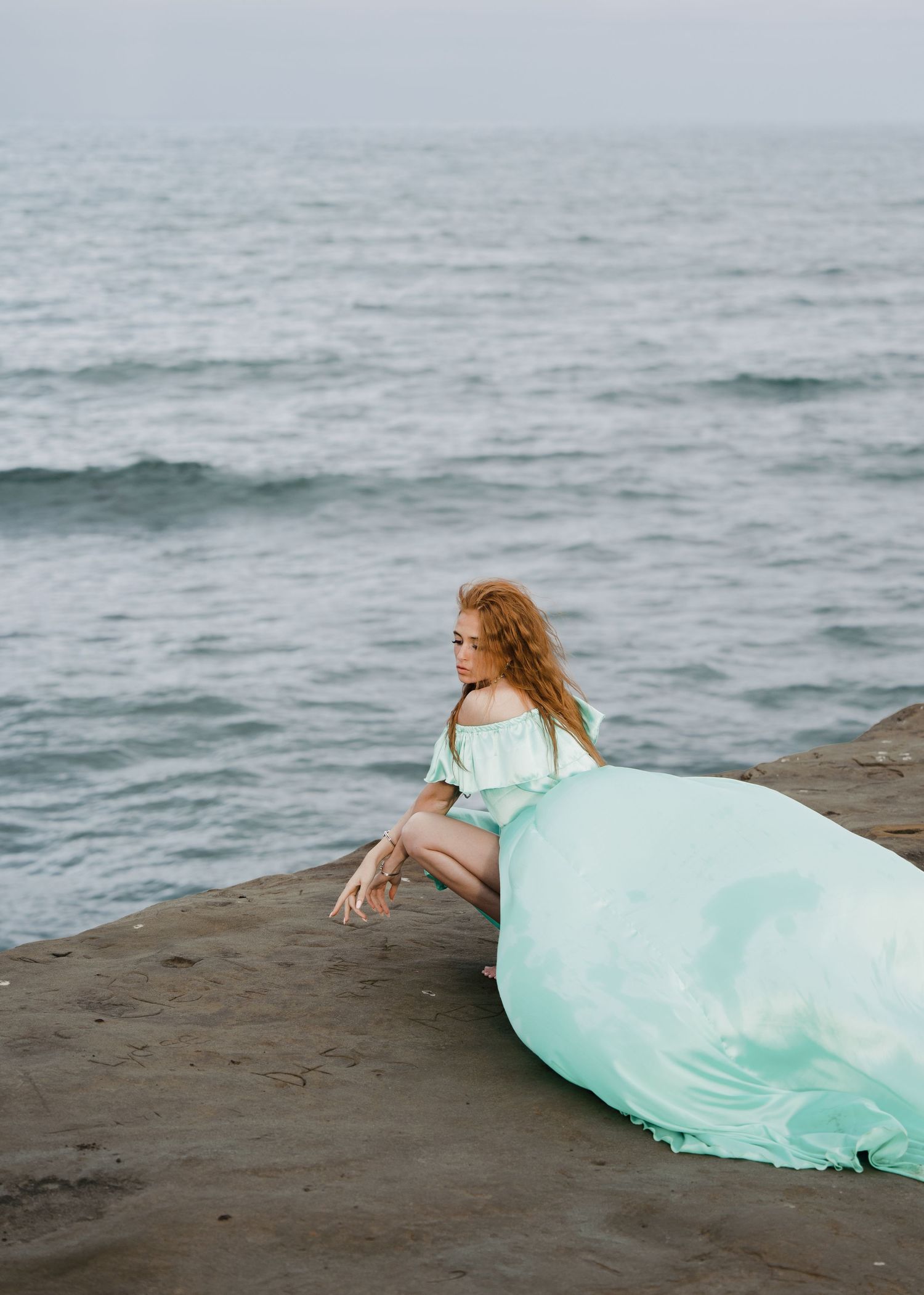 Woman in elegant mint green gown sitting on rocky coastal cliffs overlooking the sea.