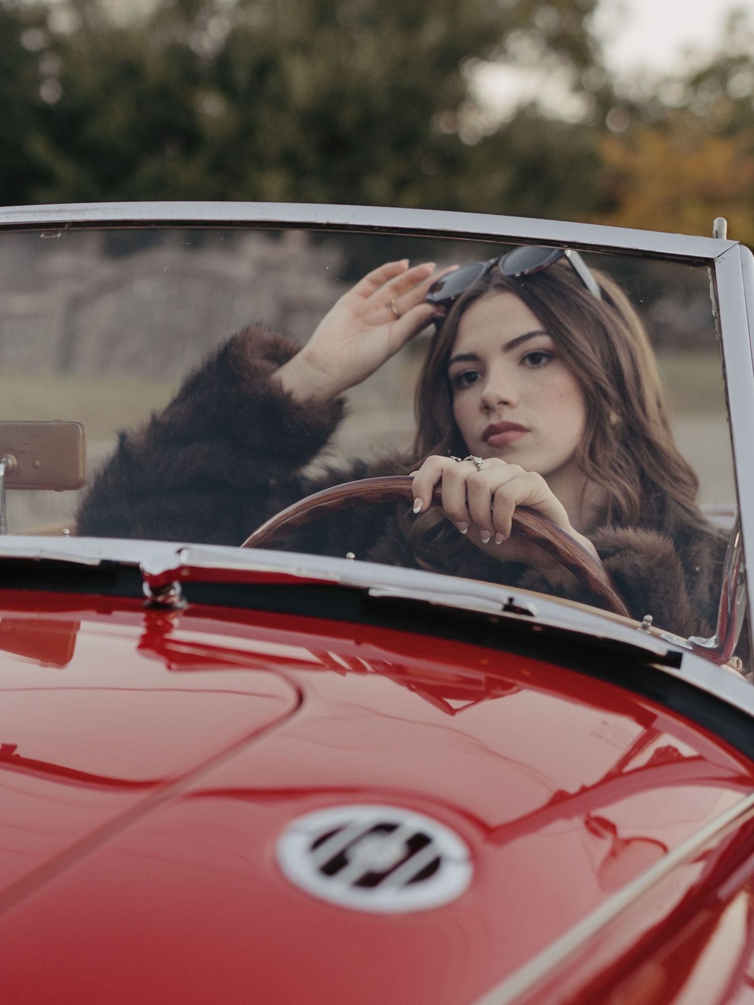 Young woman in fur coat grips steering wheel of classic red convertible sports car.