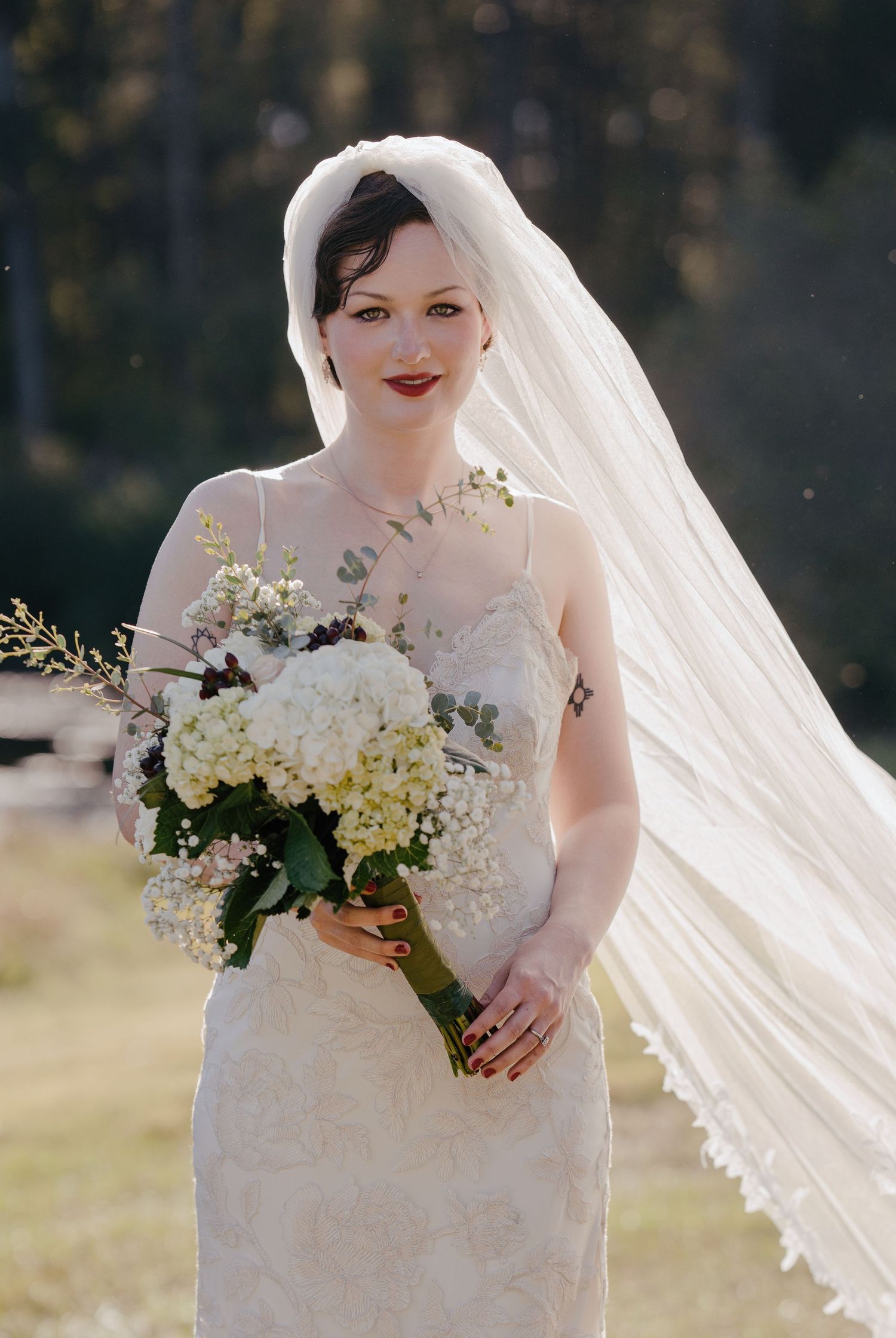 Bride in lace gown holds white hydrangea bouquet with veil flowing in the breeze outdoors.
