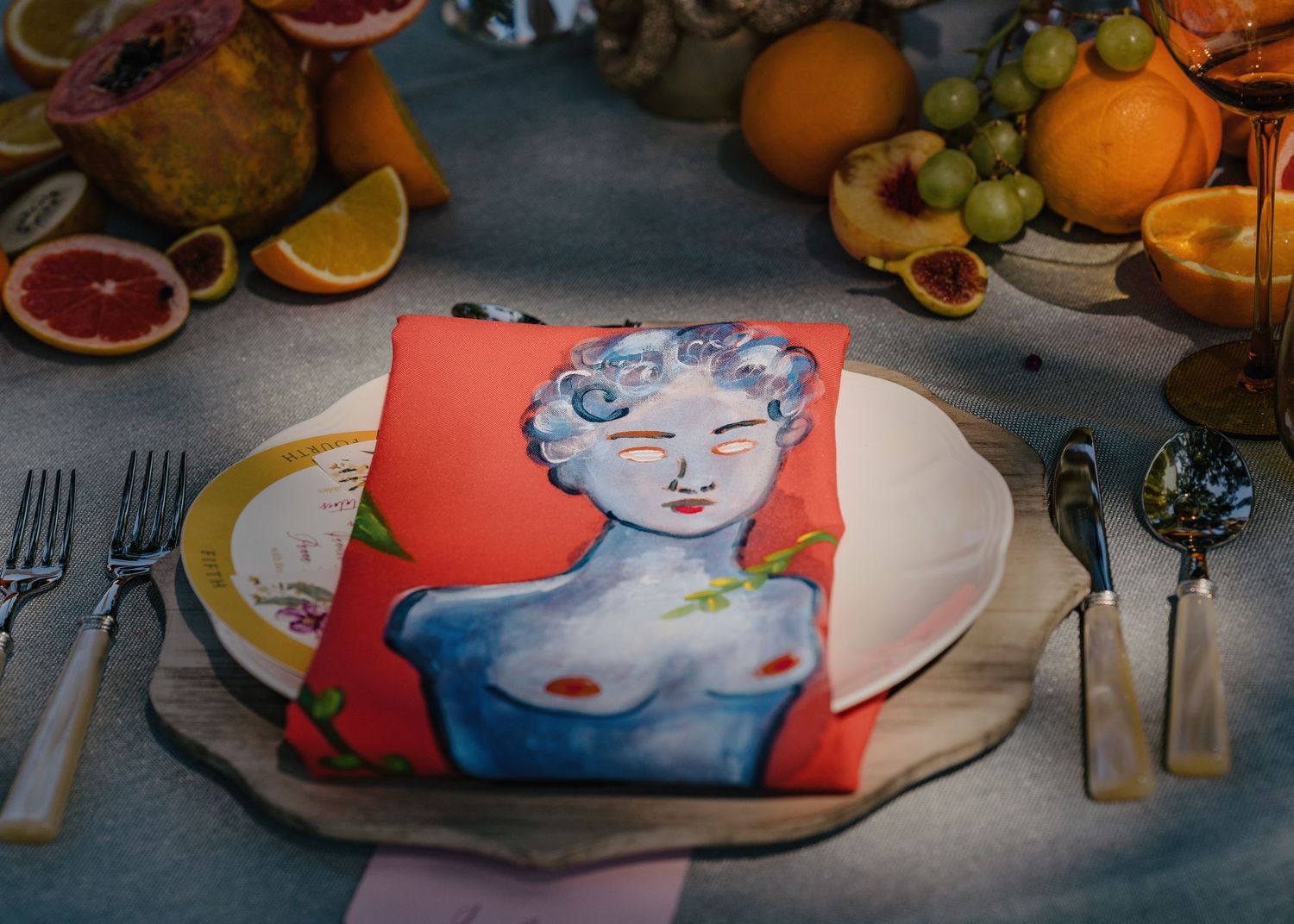 Artistic wedding place setting with painted napkin featuring female figure amid fresh tropical fruits.