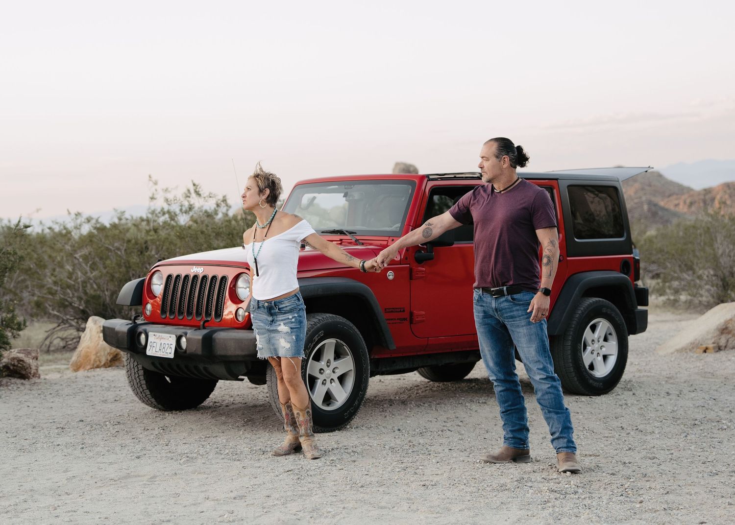 Couple standing apart beside a red Jeep Wrangler in an open desert terrain at dusk.