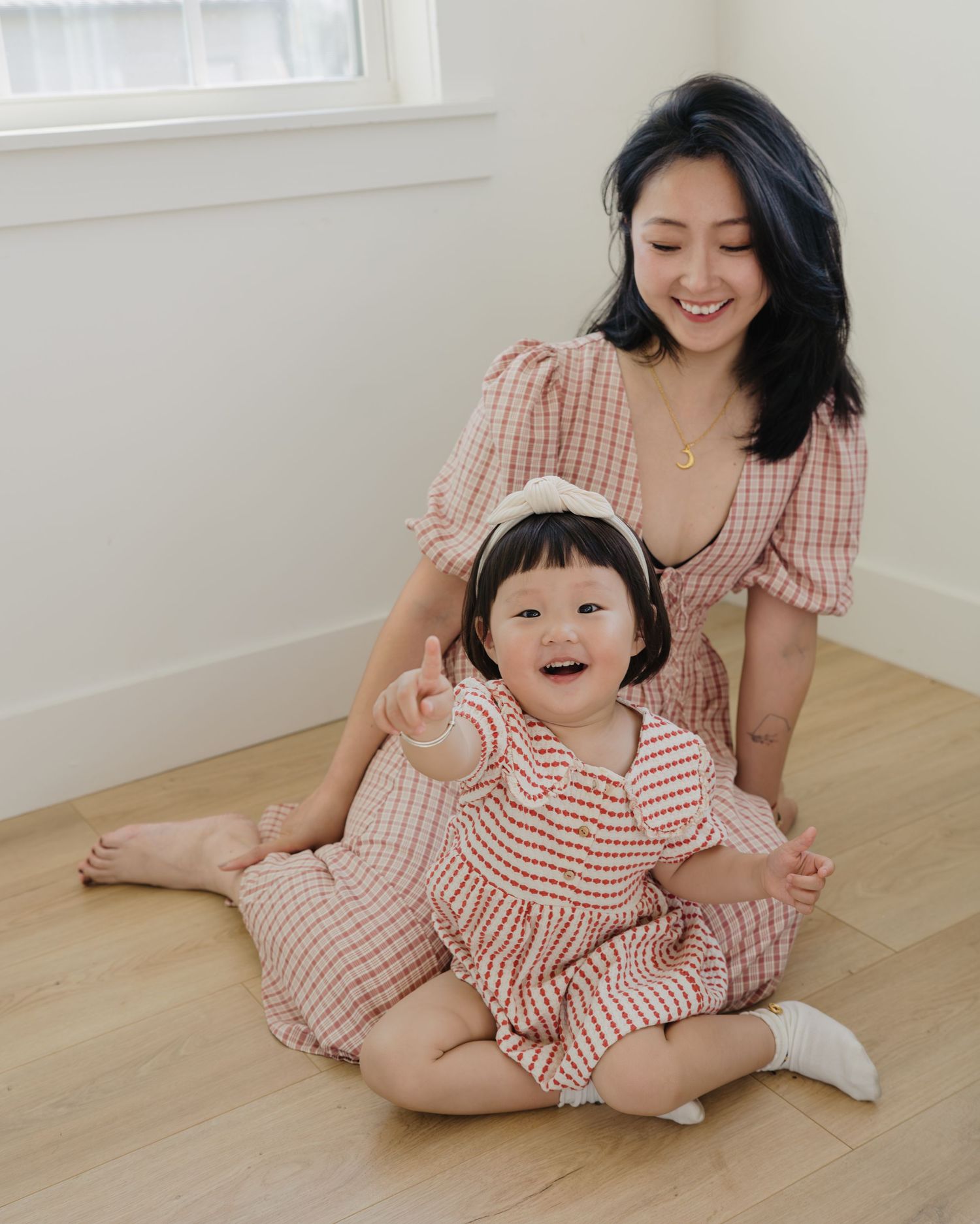 A smiling Asian mother and toddler daughter in matching pink gingham outfits sitting on a wooden floor.