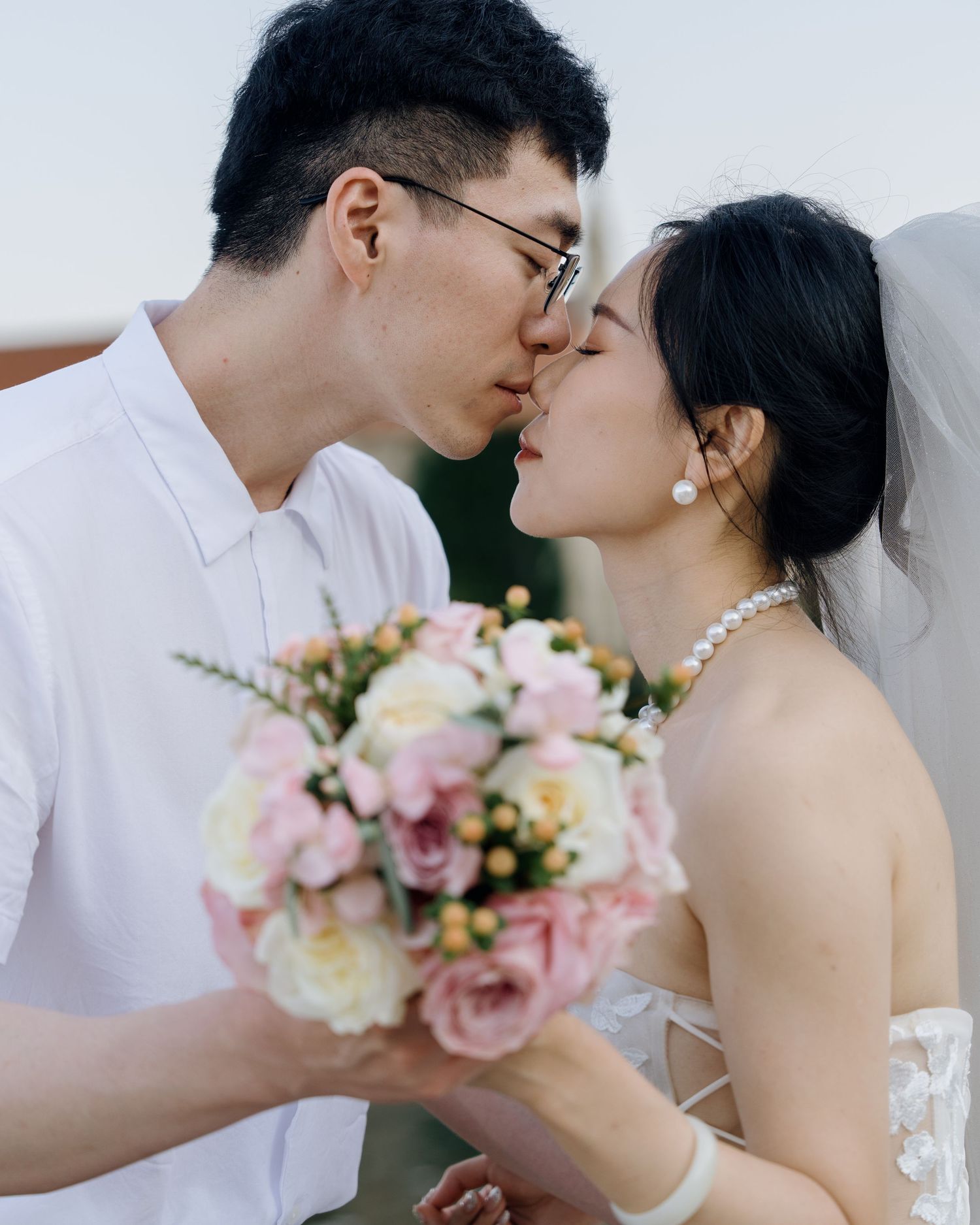 A couple shares a romantic kiss, the bride holding a pink and white bouquet while wearing a veil and pearl necklace.