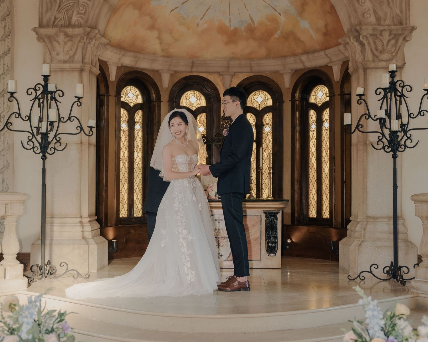 Bride and groom stand at the altar of an elegant chapel with arched stained glass windows.
