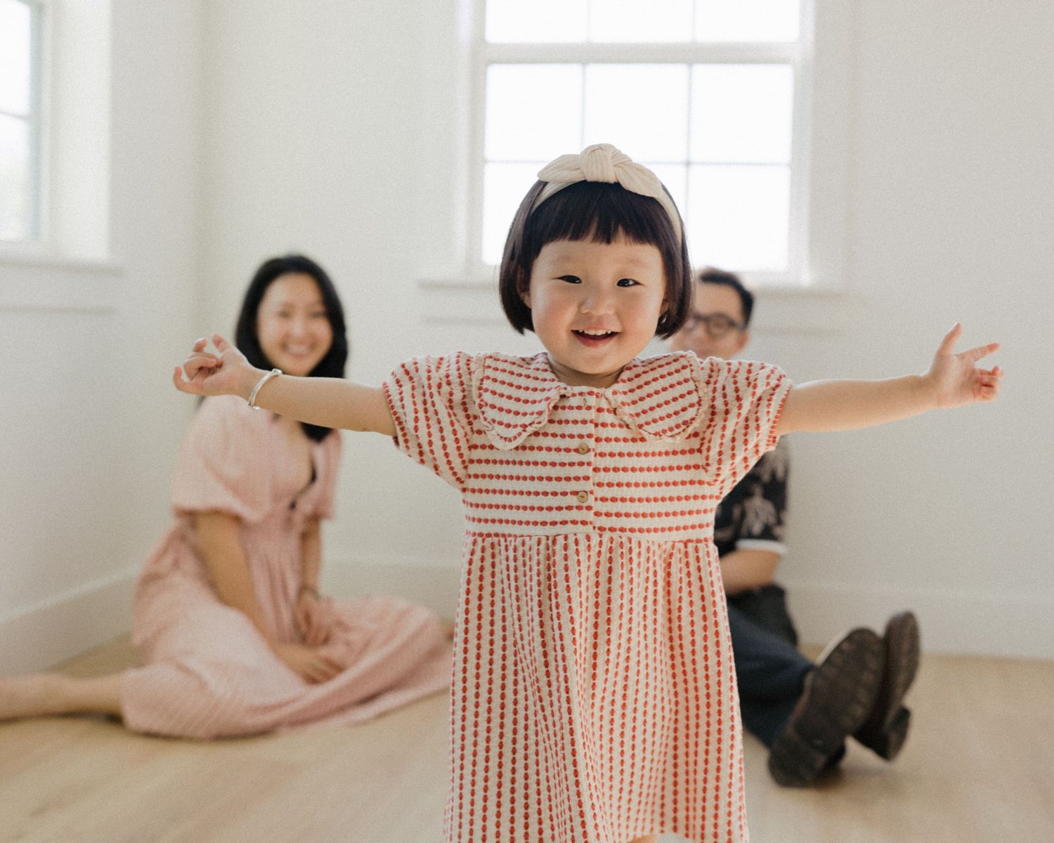 A joyful toddler in a pink gingham dress spreads her arms wide while her parents sit smiling behind her.