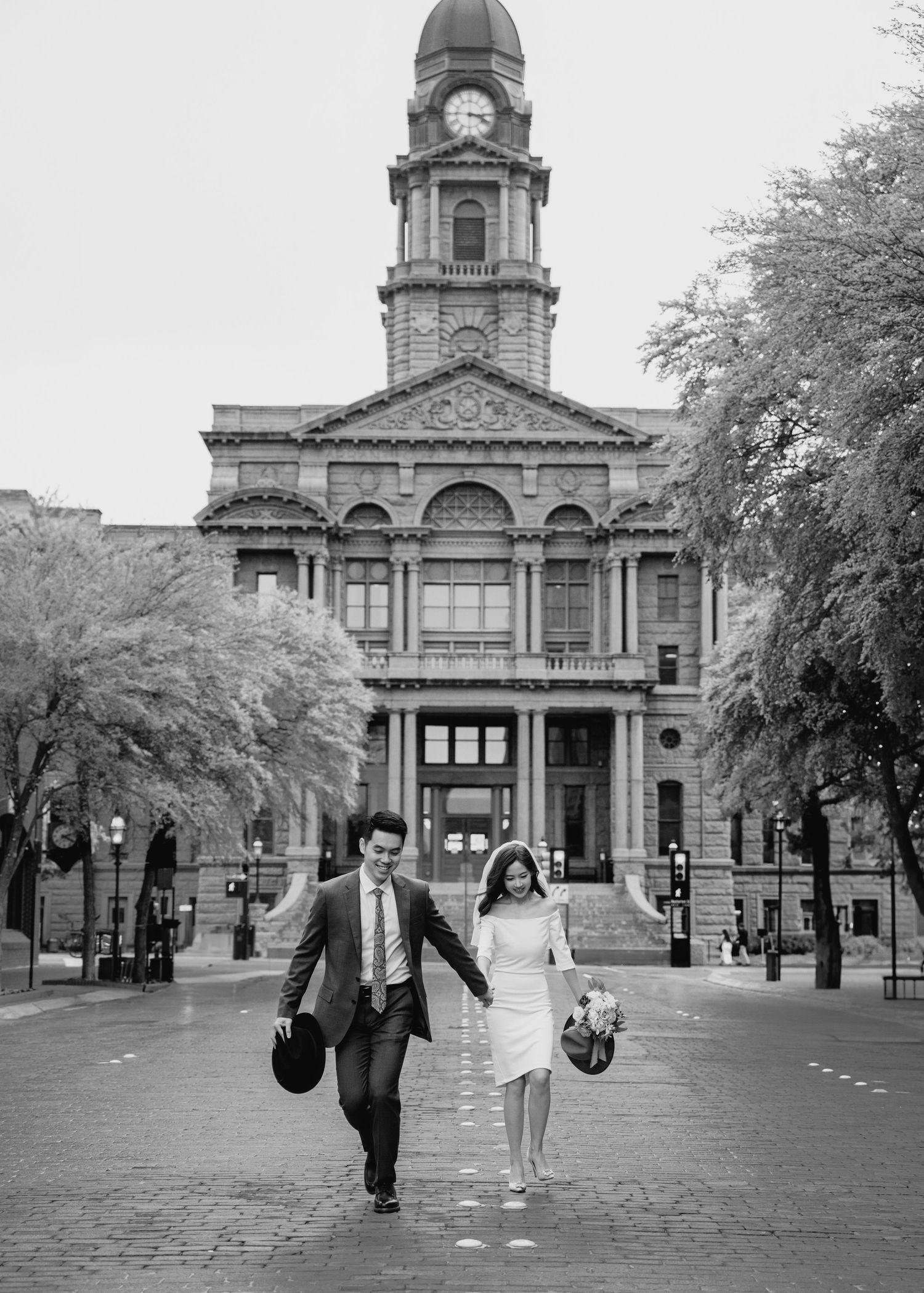 Happy couple runs hand-in-hand in front of the Tarrant County historic courthouse in black and white photo.