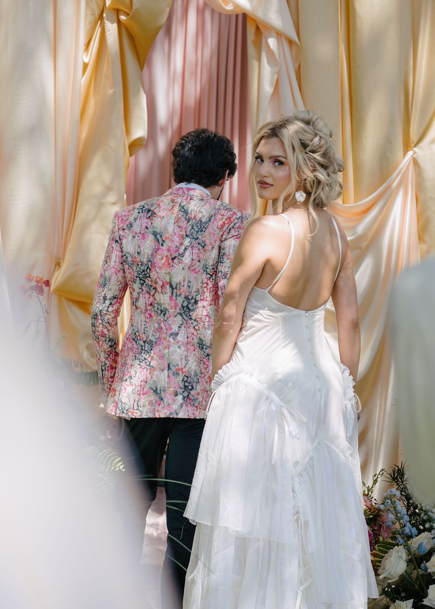 Bride in backless white wedding dress walks with groom in floral blazer under draped pink and gold arch.