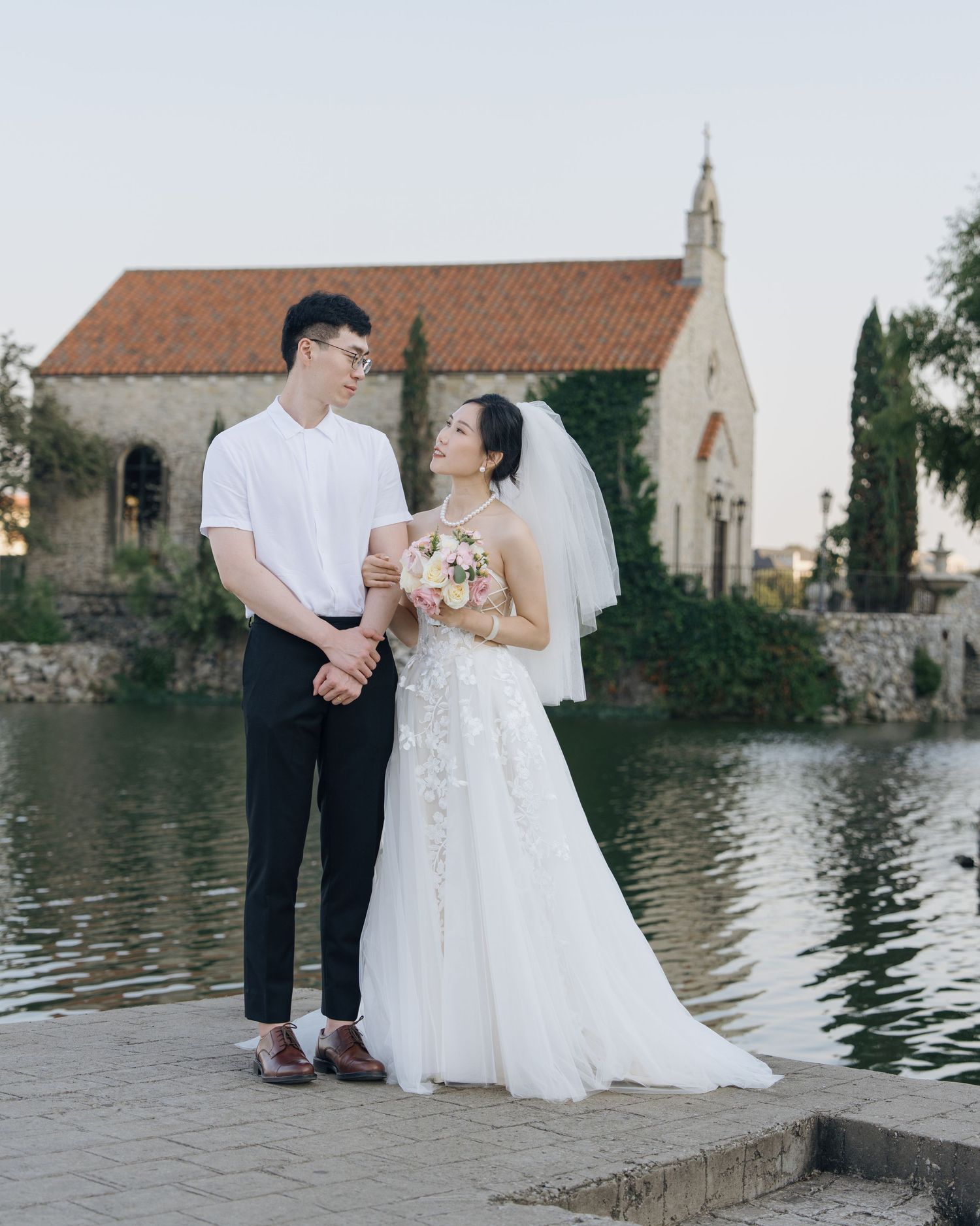 Elegant bride and groom standing by a lake with a historic stone chapel backdrop at sunset.